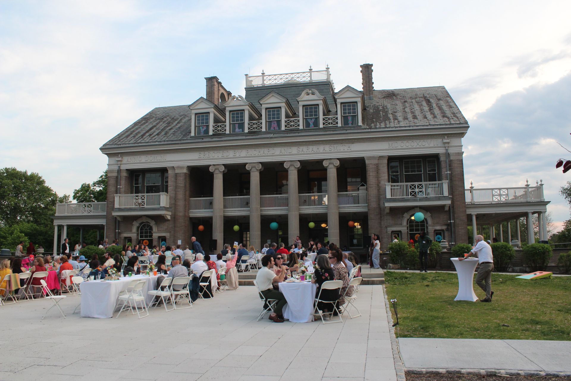 Crowd gathered outside a large historic mansion for an outdoor event with tables and chairs on the lawn
