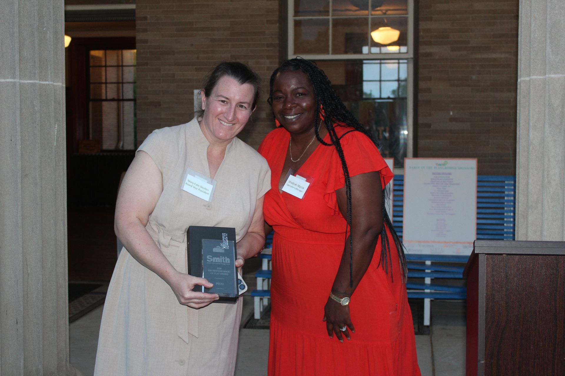 Two people posing indoors, one holding an award plaque, in front of a brick wall and blue bench