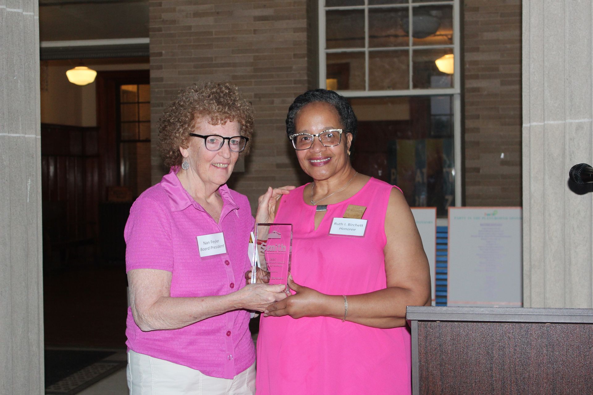 Two women in pink shirts smile and shake hands indoors at a reception desk.