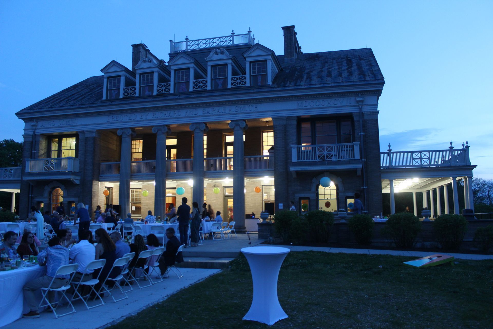 Evening outdoor dinner event at a lit historic mansion with guests seated on the lawn