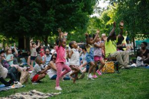 Children dancing in a park while an audience sits on the grass watching.