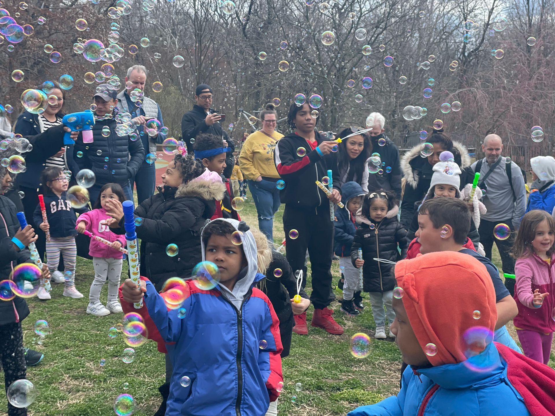 Crowd of people outdoors blowing soap bubbles, with children bundled in coats in a park