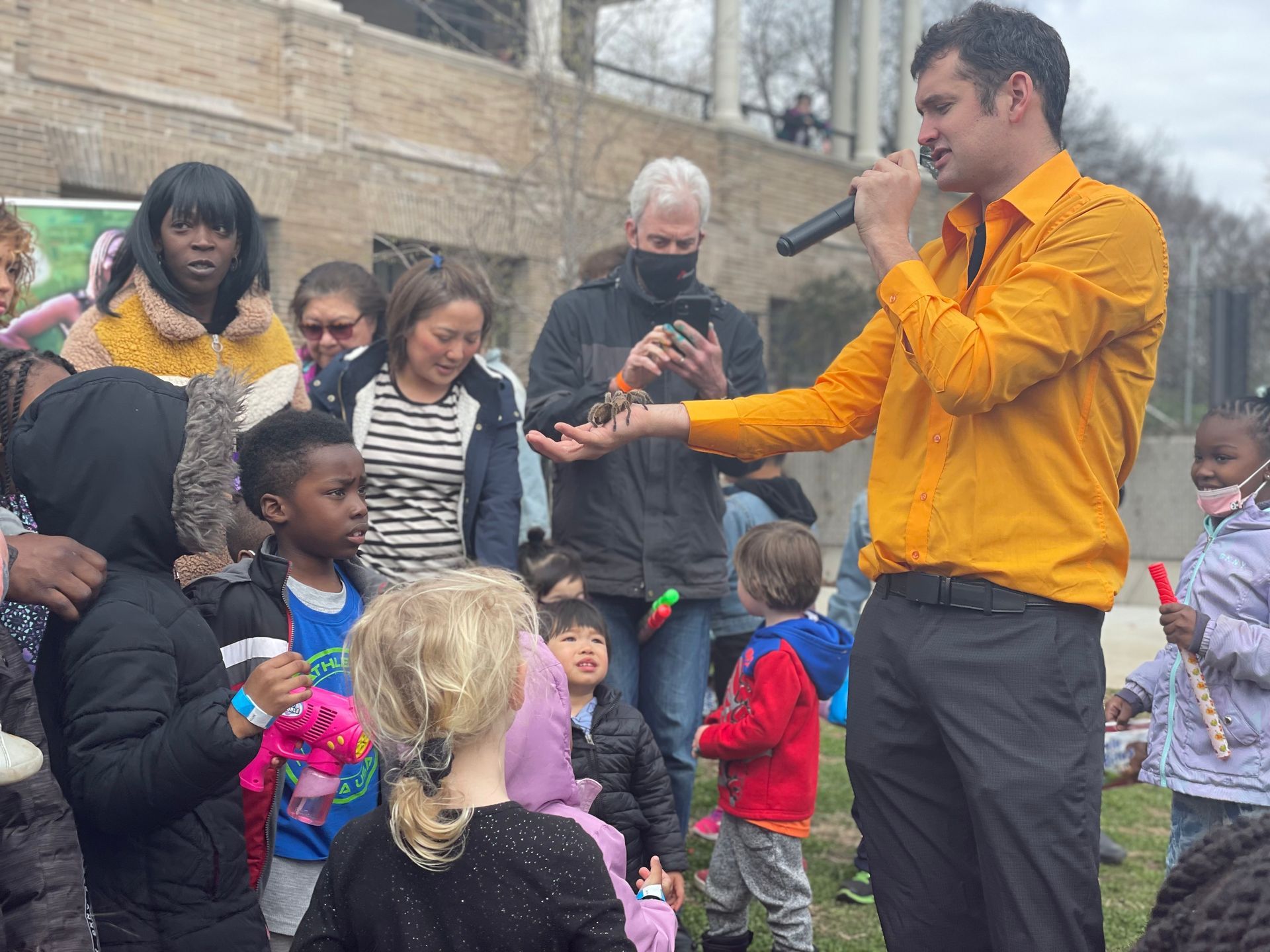 Man in a bright orange jacket speaking to a small crowd outdoors, with children gathered around him