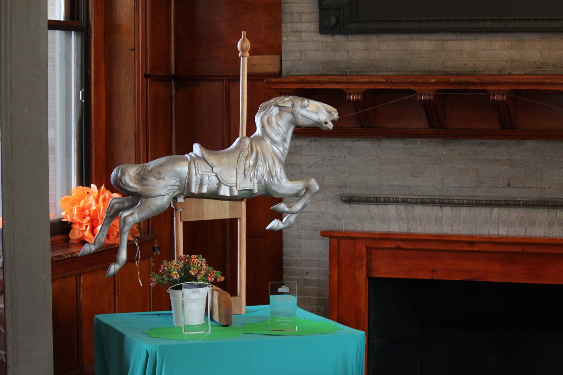 Silver rocking horse sculpture with orange tail on a turquoise table beside a fireplace.