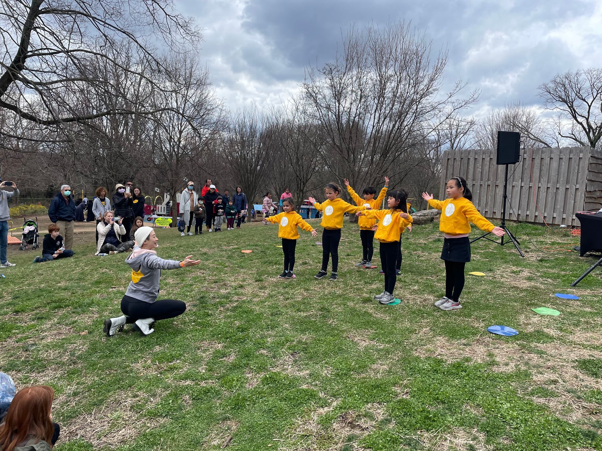 Children in yellow shirts dancing outdoors while a crouching instructor leads them in a park