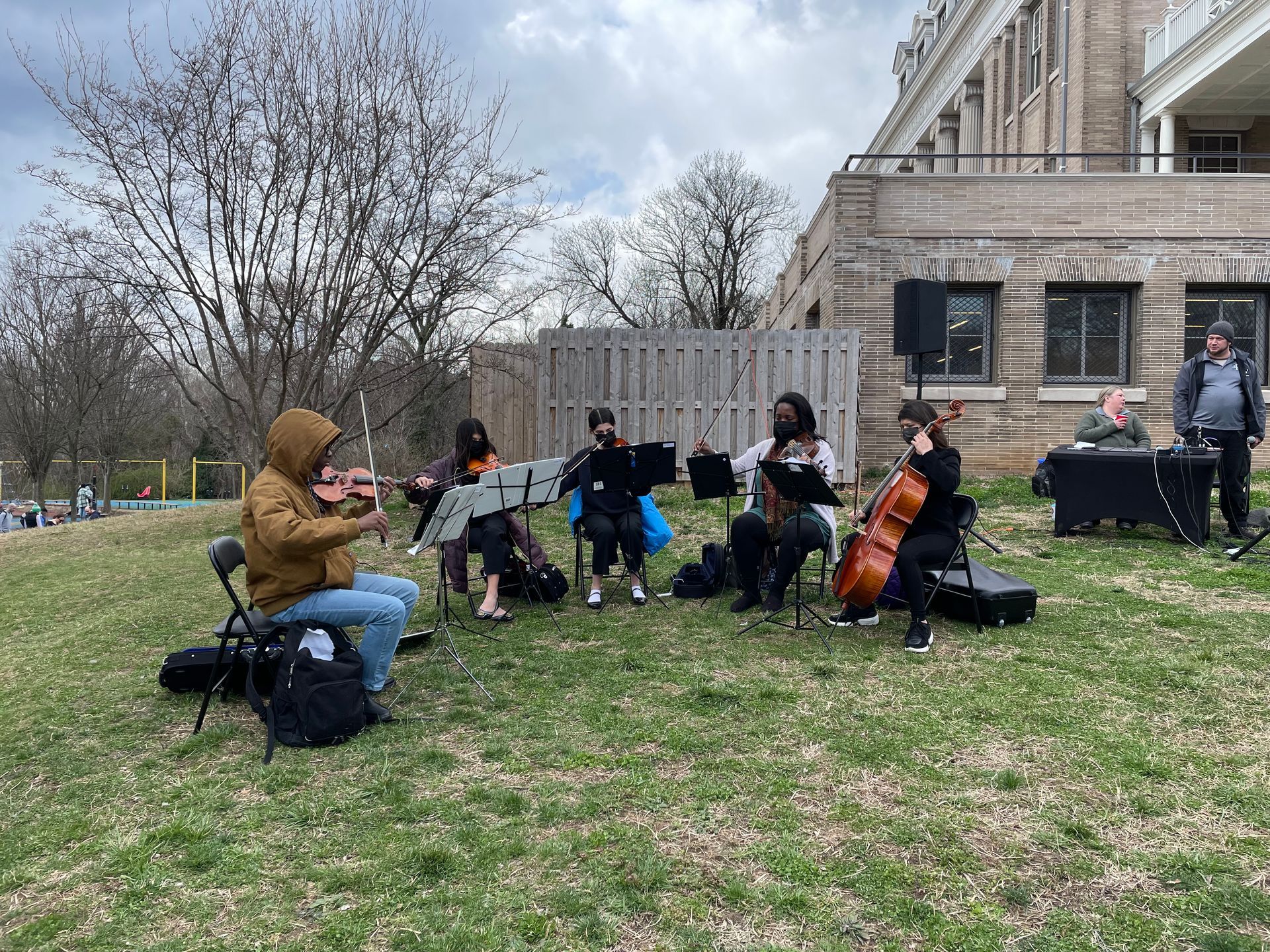 Small outdoor string ensemble performing on a grassy lawn beside a stone building in early spring