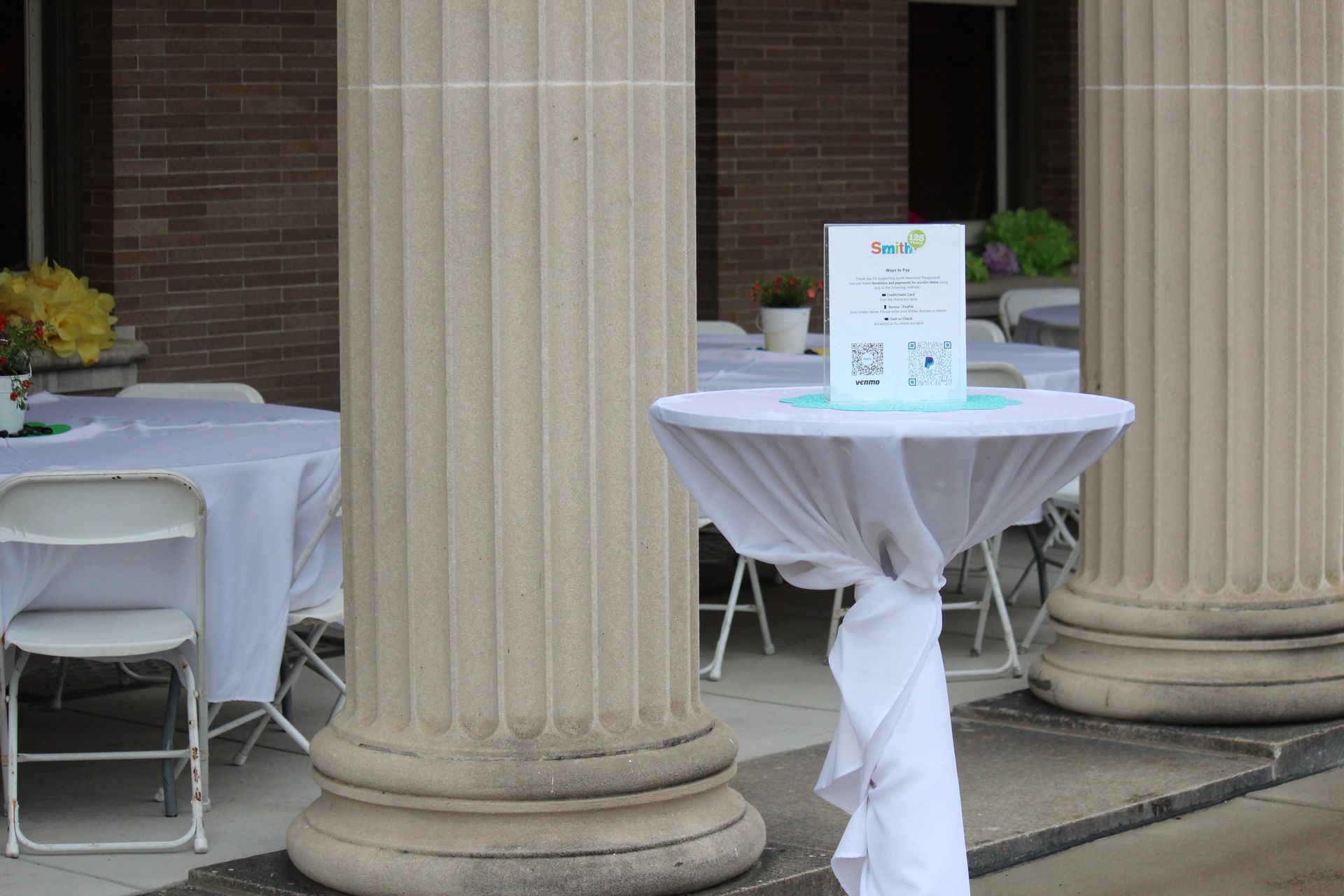 Decorated outdoor tables between large stone columns, with white tablecloths and a sign on one table