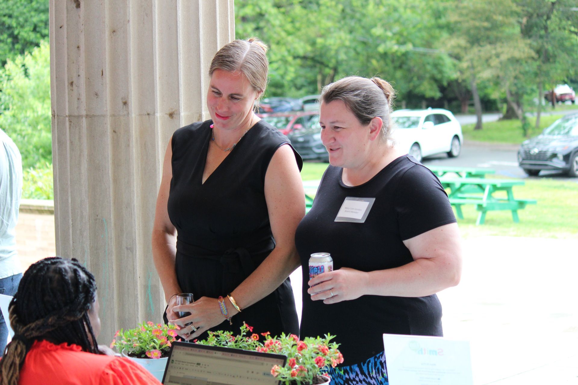 Two women stand behind a flower display at an outdoor booth, one holding a drink.