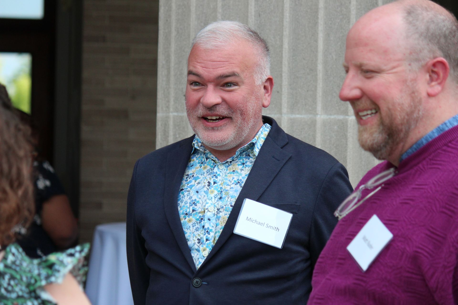 Two men smiling and talking at an outdoor event, wearing name tags and business-casual clothing.