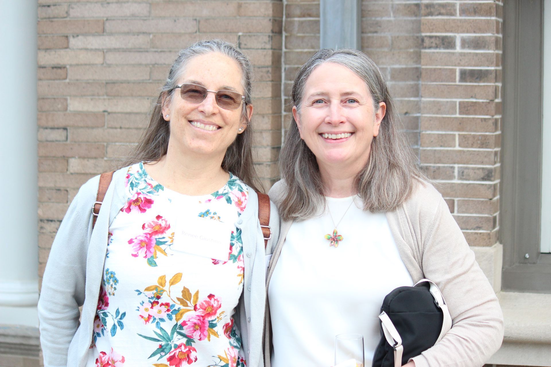 Two smiling women standing outdoors in front of a brick wall, one holding a black jacket