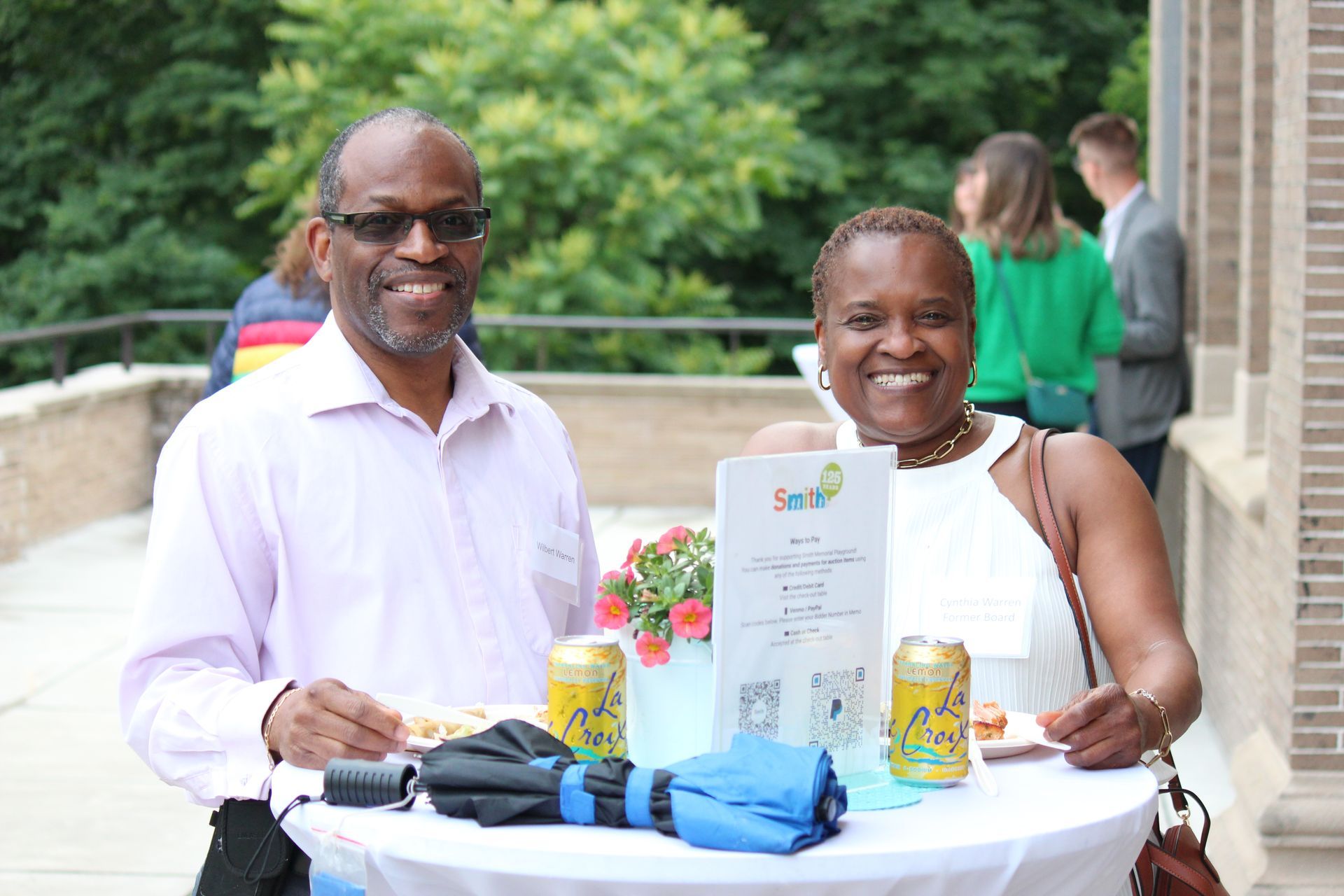 Two smiling people at a table with a certificate, flowers, and gifts outdoors
