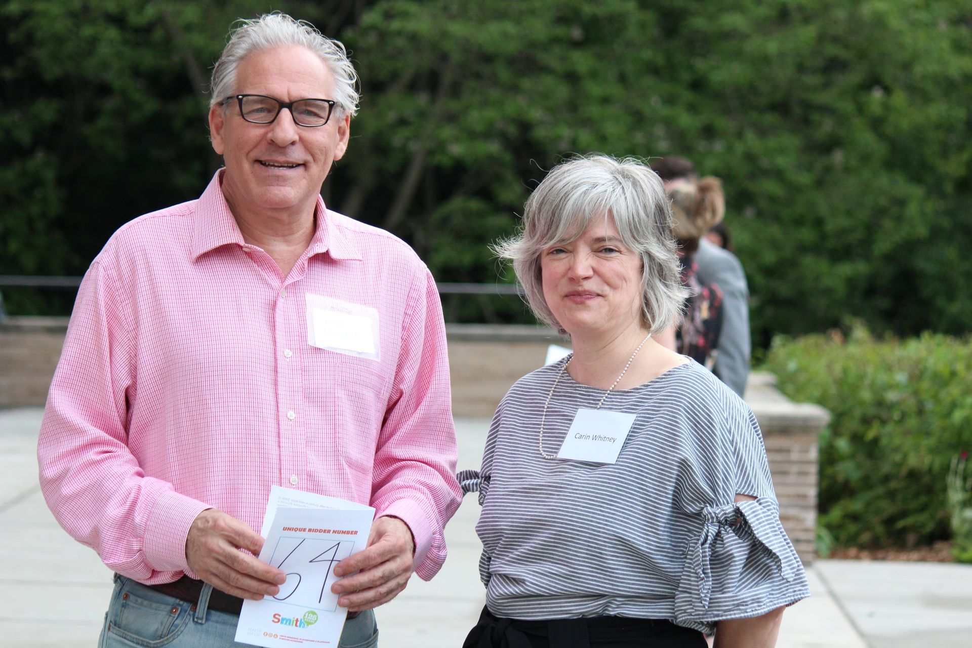 Two people standing outdoors, one holding papers, with a green backdrop.