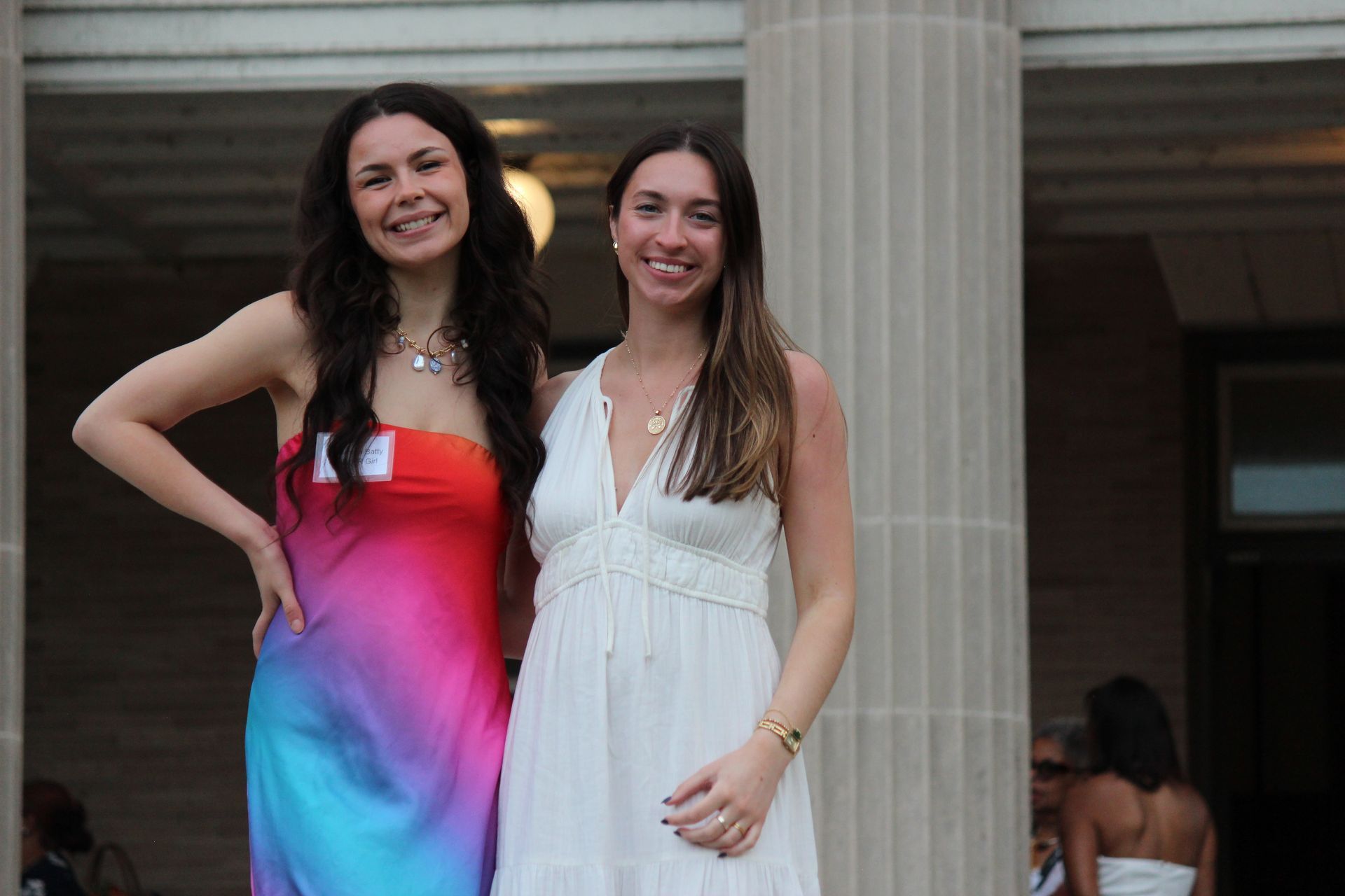 Two women posing together, one in a colorful dress and one in a white dress, standing by a column