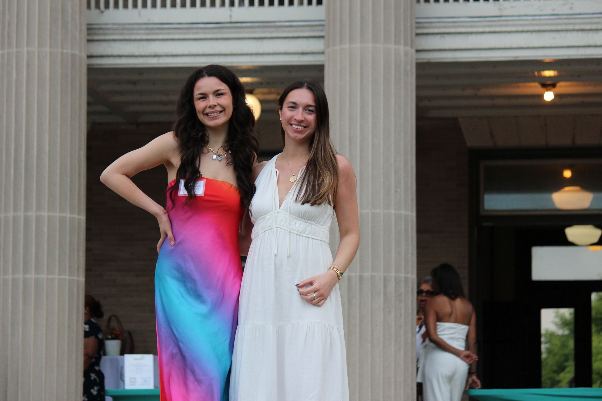 Two women pose in front of a building, one in a pink-blue dress and one in a white dress.