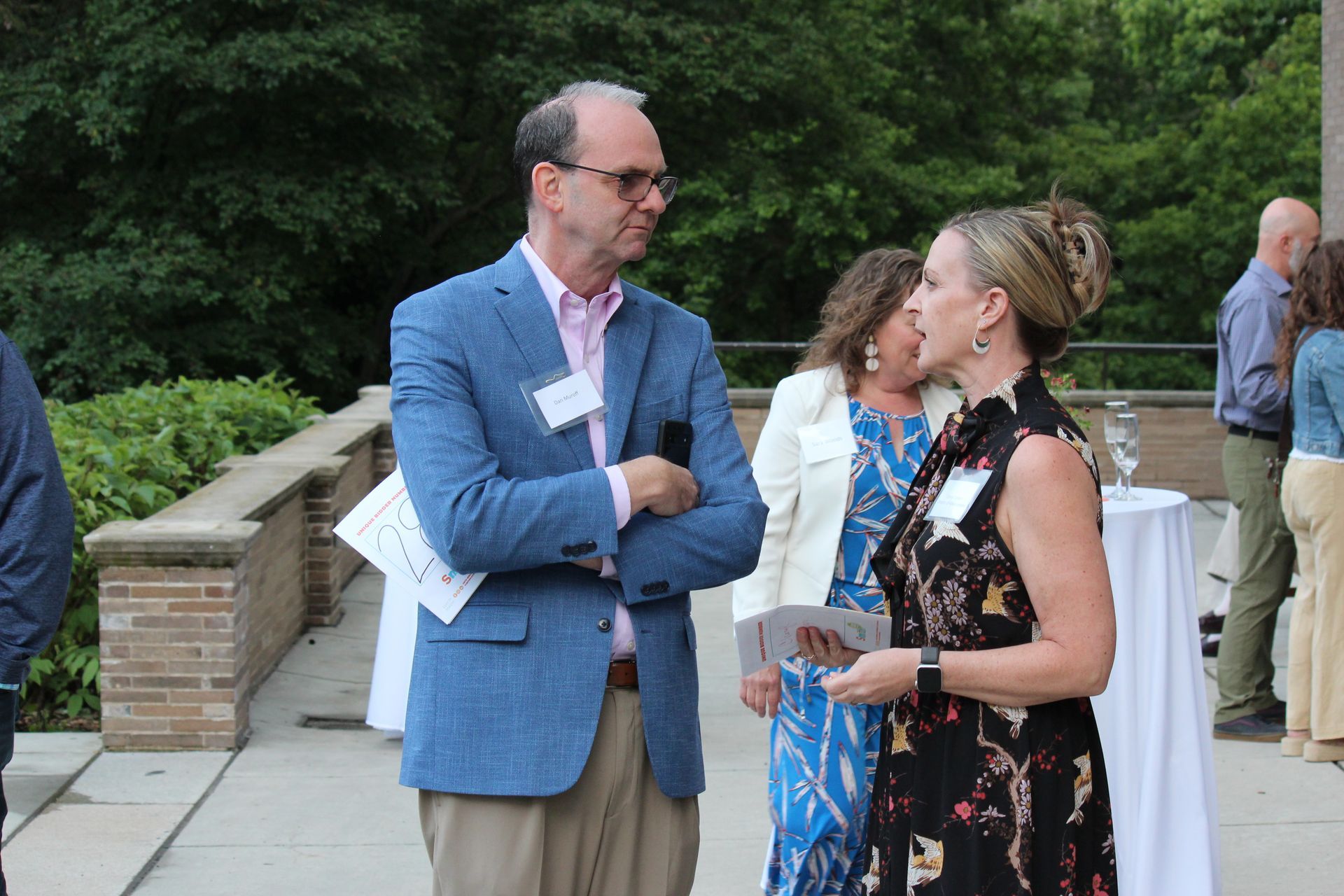 Two people chatting outdoors at a garden gathering, one in a blue blazer and one in a floral dress.