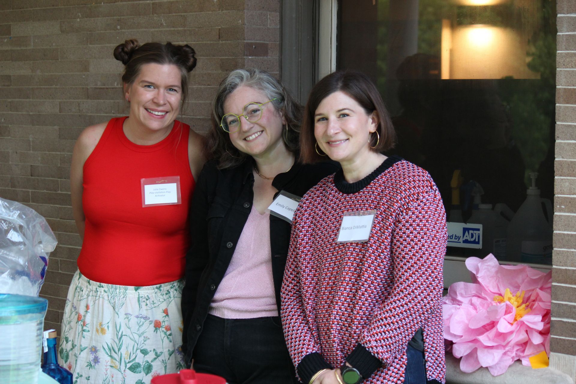 Three people smiling at a table with pink flowers and event name tags indoors.