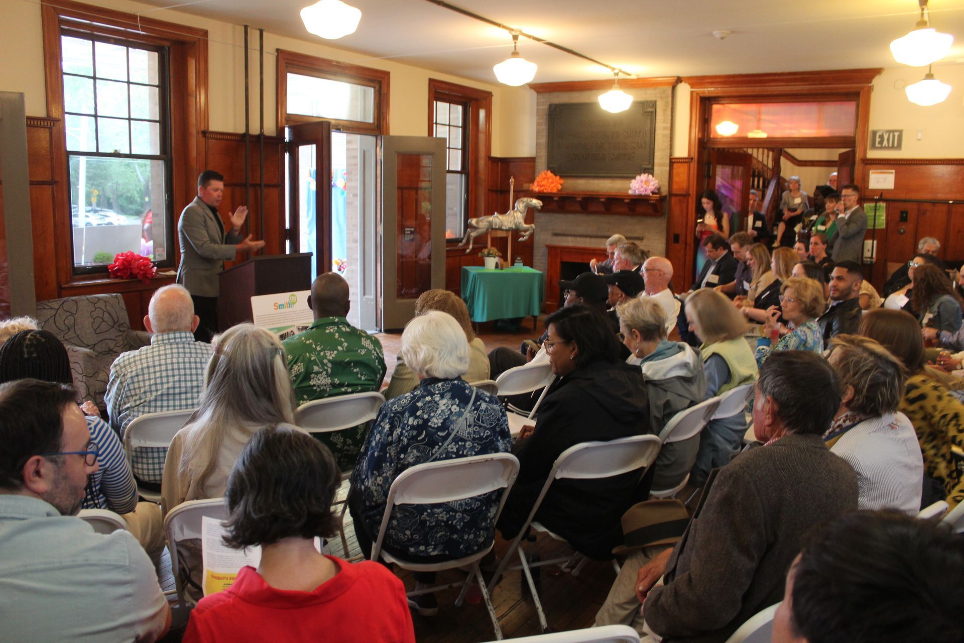 Audience seated in a living room for a community talk, speaker standing near the window
