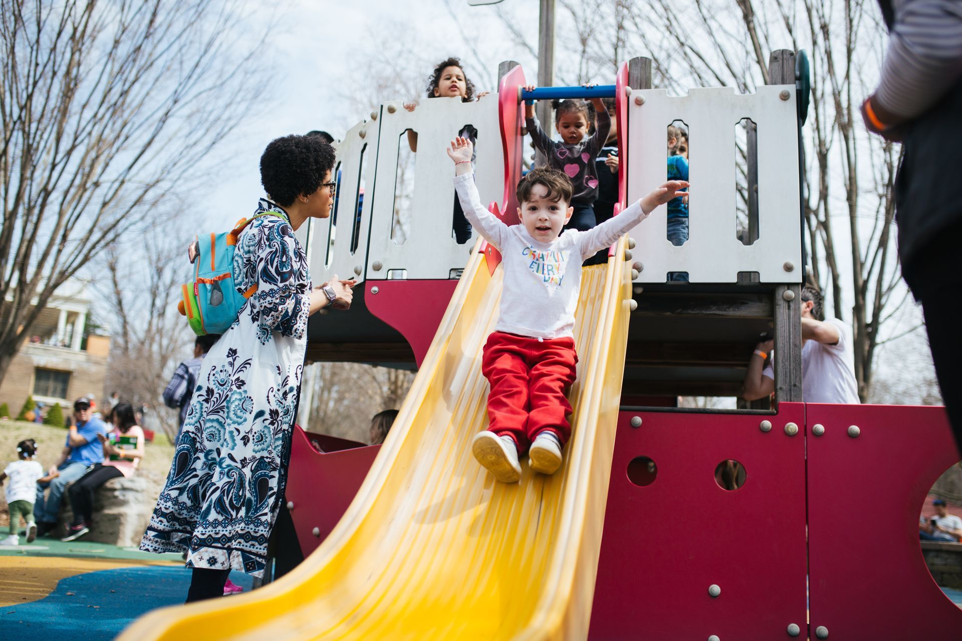 Child sliding down a yellow slide at a playground, with other children and adults nearby