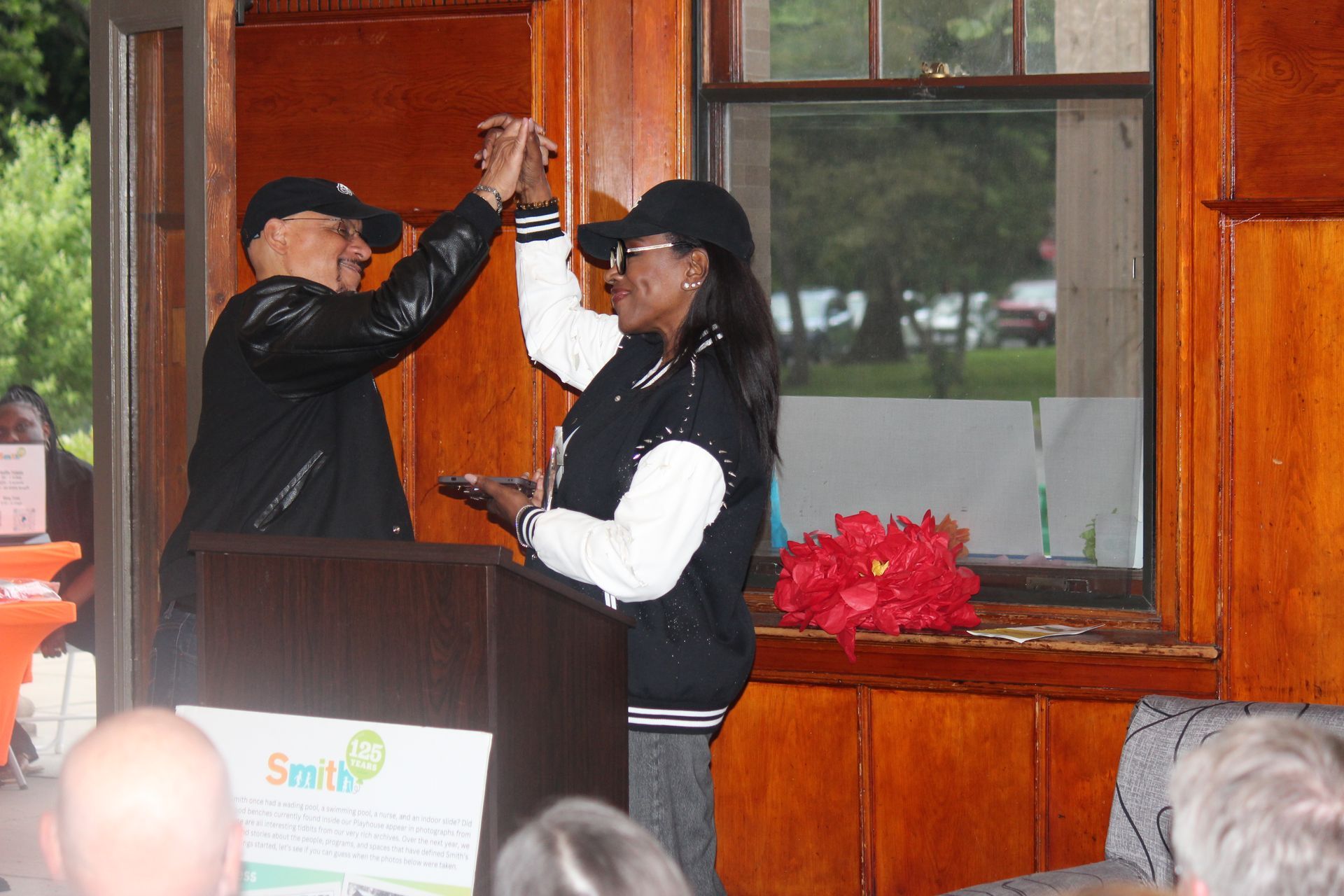 Two people high-five beside a podium in a wood-paneled room, with flowers on the windowsill.
