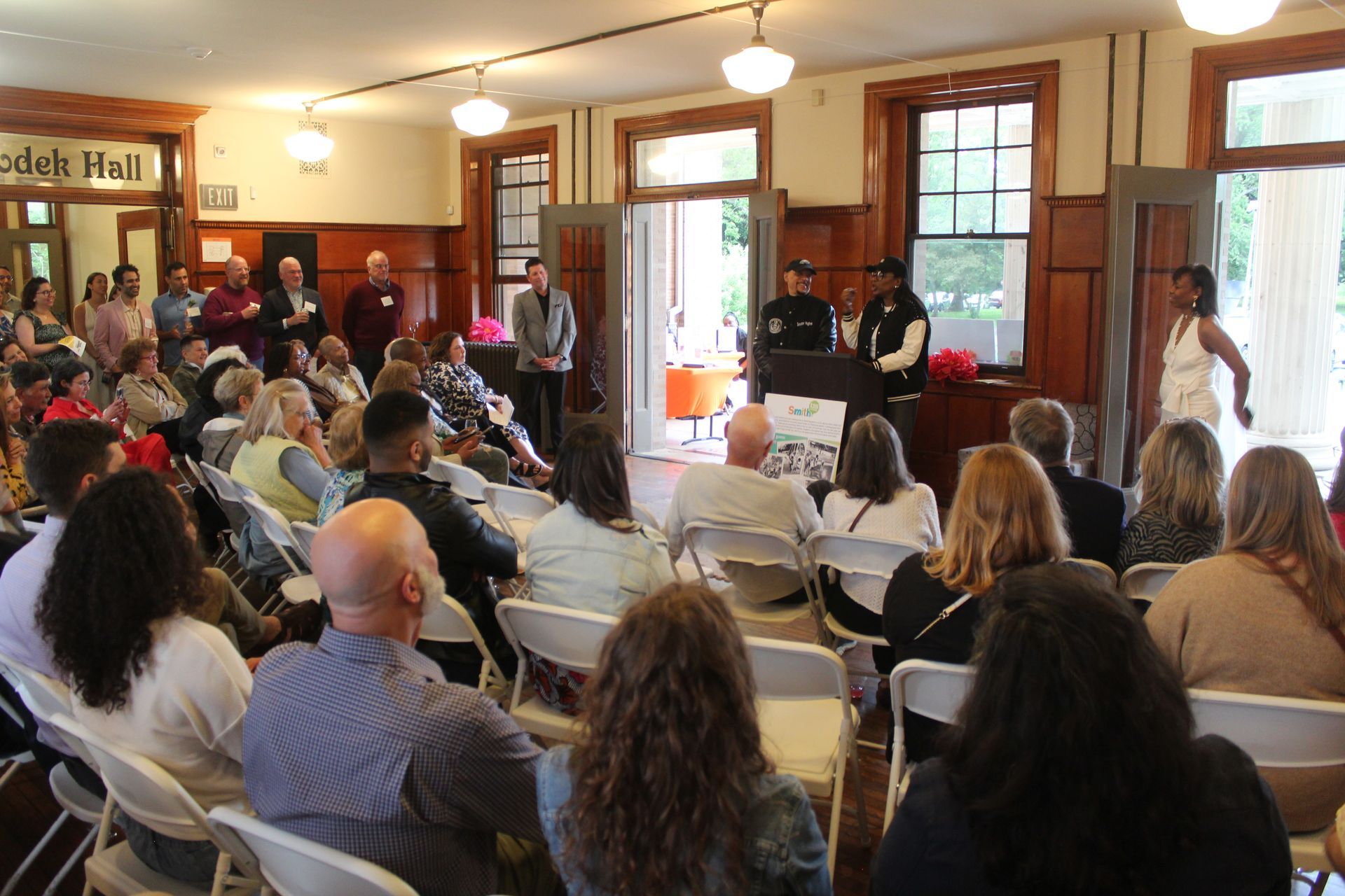 Audience seated in a bright room, watching a speaker near the doorway at a community event.