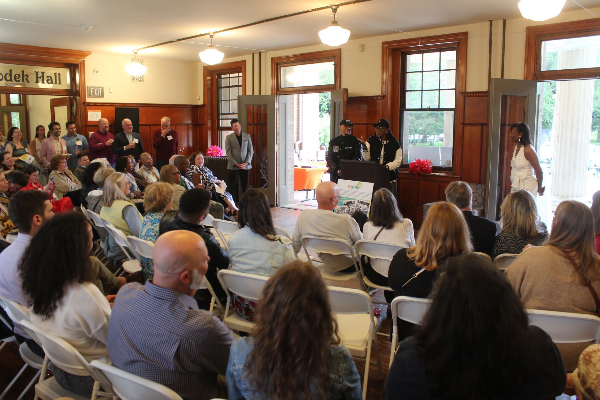 Audience seated in a cozy room watching a speaker at the front near tall windows