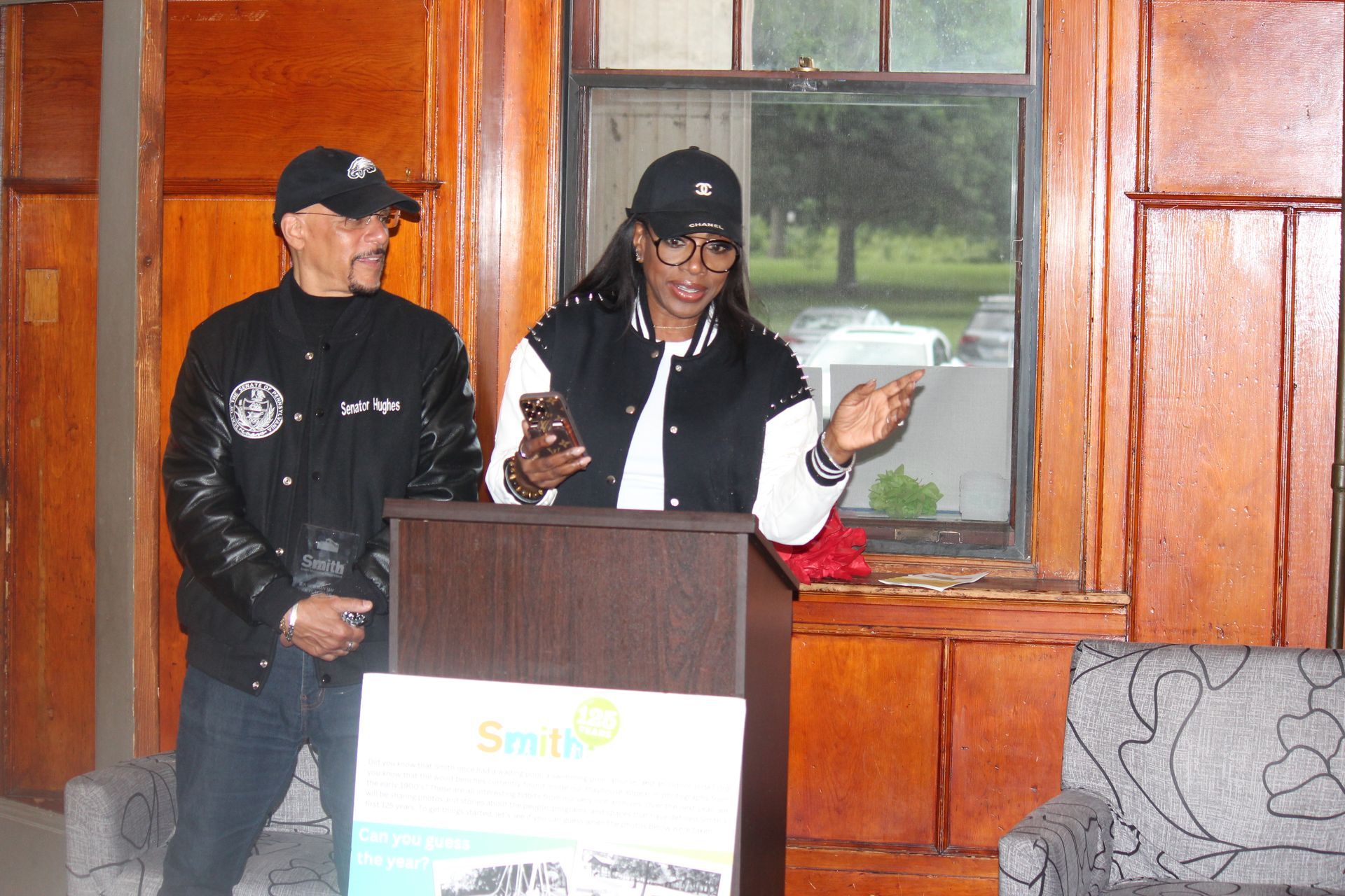 Two people standing at a podium indoors, speaking into microphones in front of wood-paneled walls.