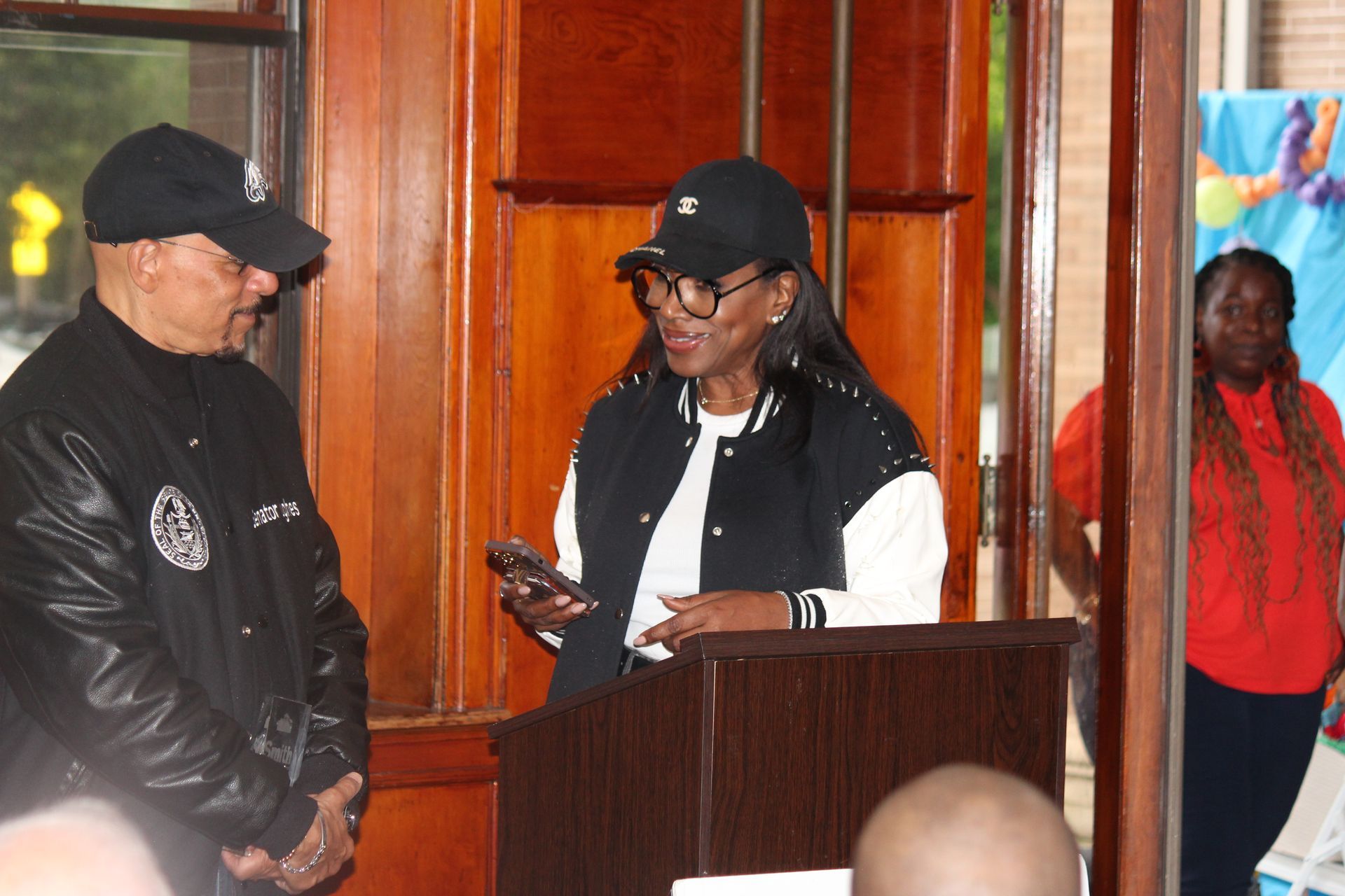 Two people stand at a podium indoors, one speaking while holding a phone and the other listens.