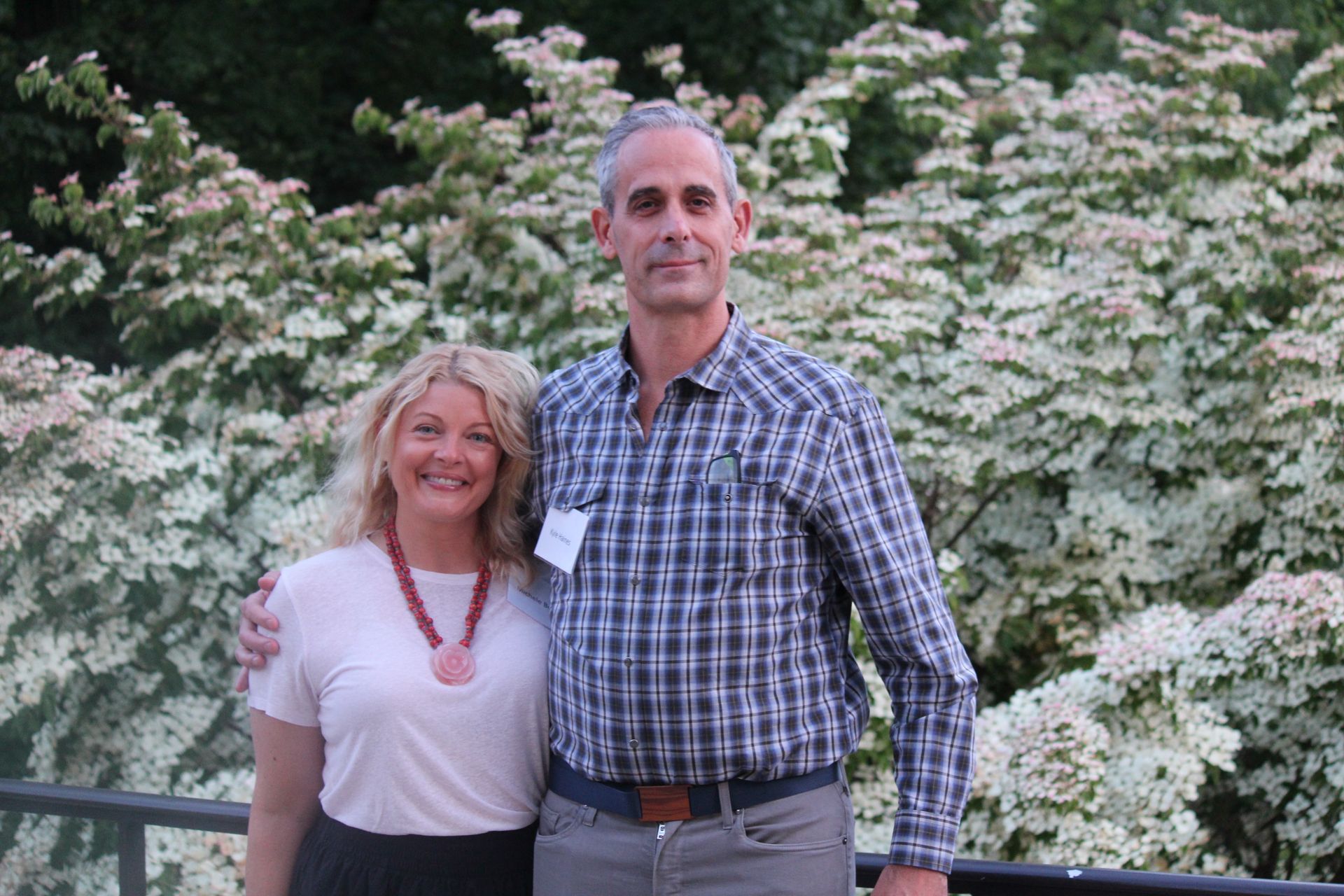 Two people standing outdoors in front of blooming white flowers, smiling at the camera.