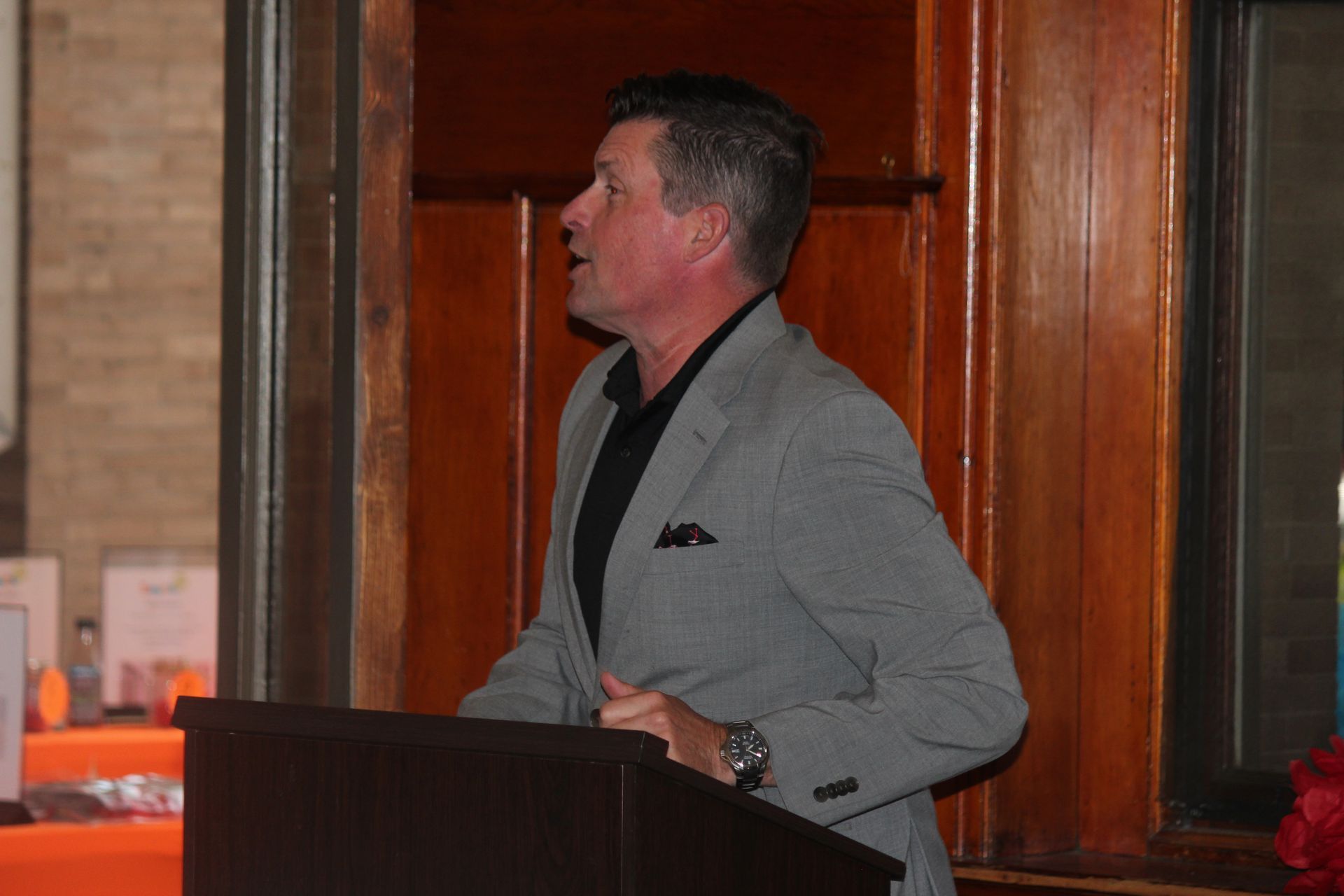 Man in a gray suit speaking at a podium indoors, side profile, in a wood-paneled room