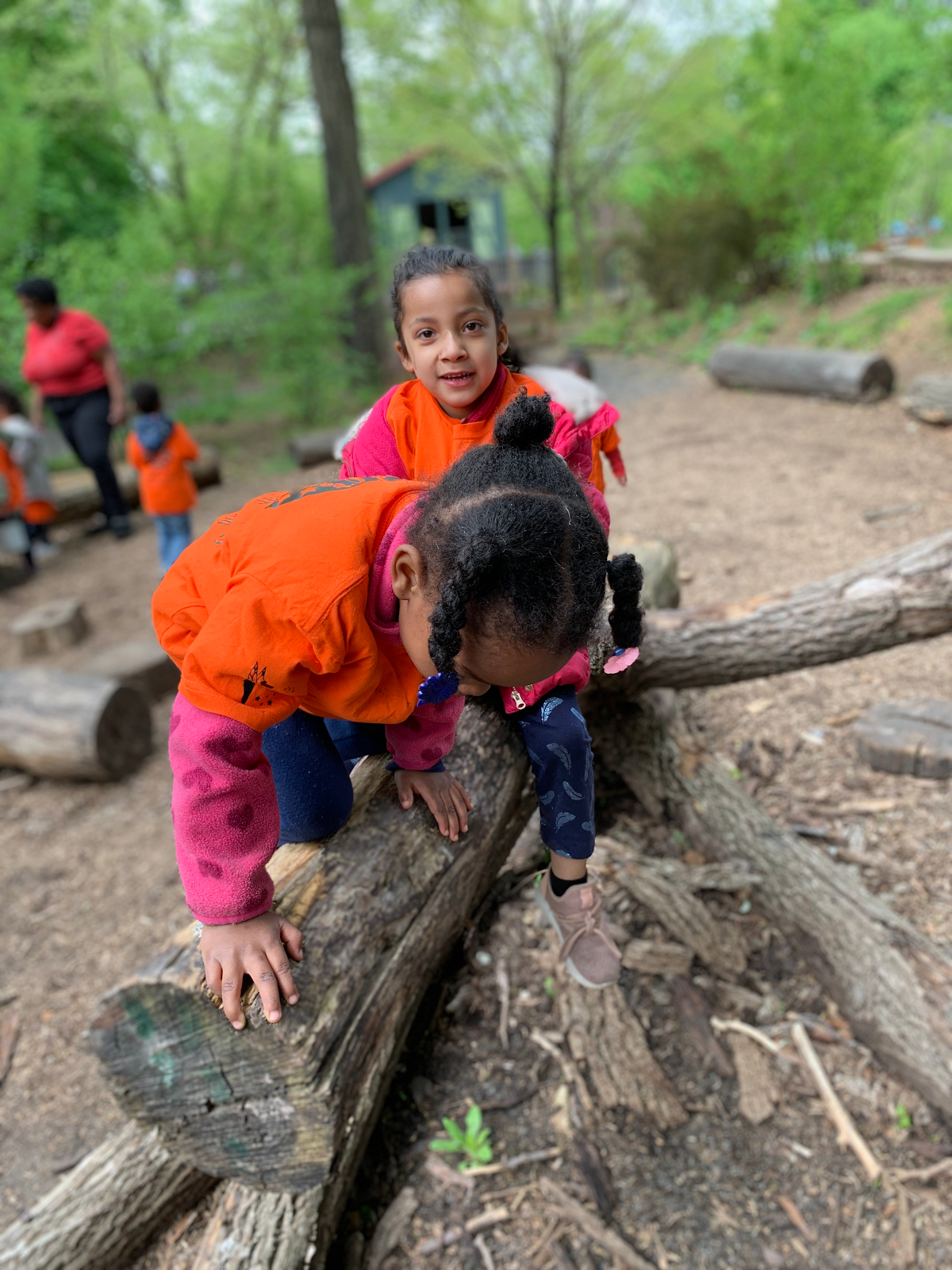 Two children in orange shirts climb over logs in a wooded outdoor play area.