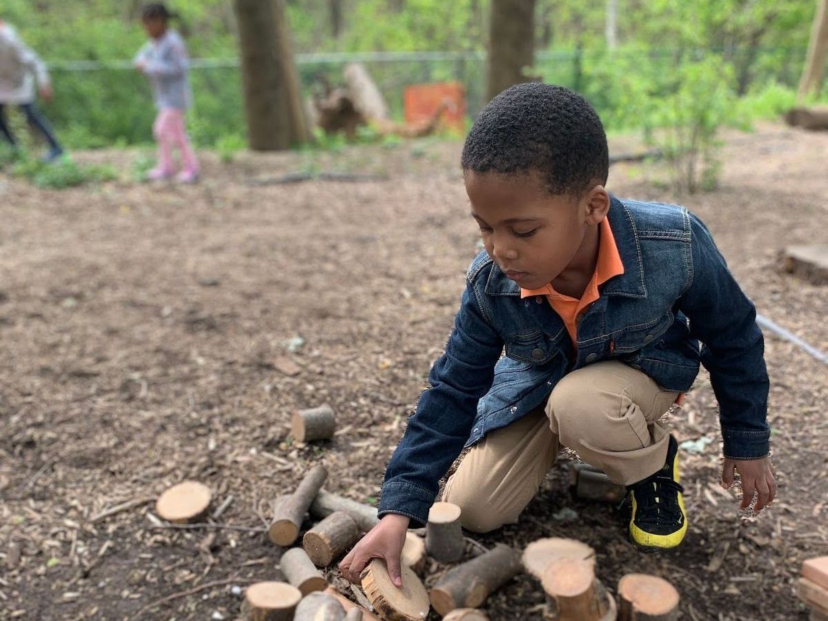 Child crouching outdoors, arranging sticks and wood rounds beside a small pile in a wooded area.