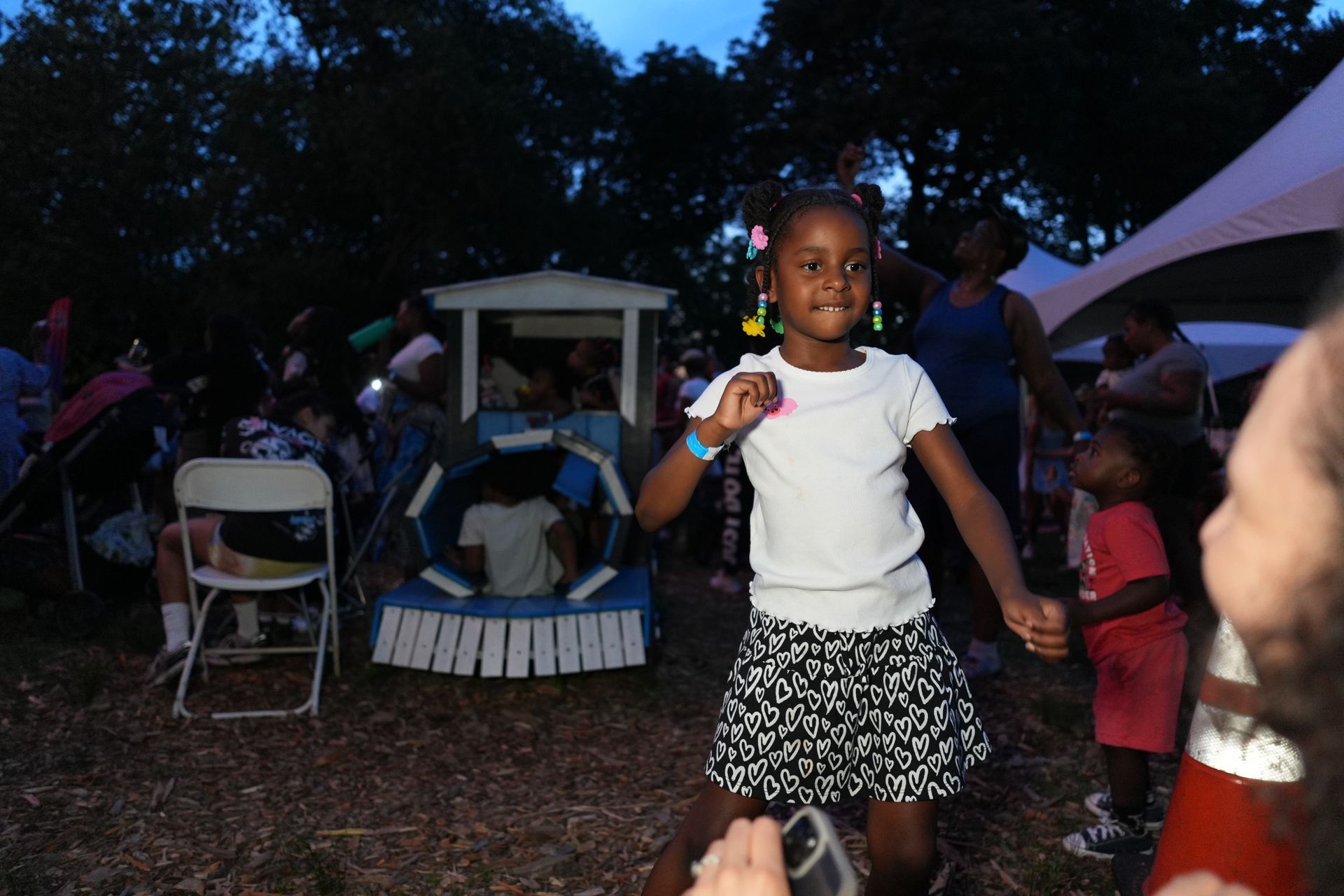 Child in white shirt and patterned skirt standing outdoors at dusk near seated people and a tent.