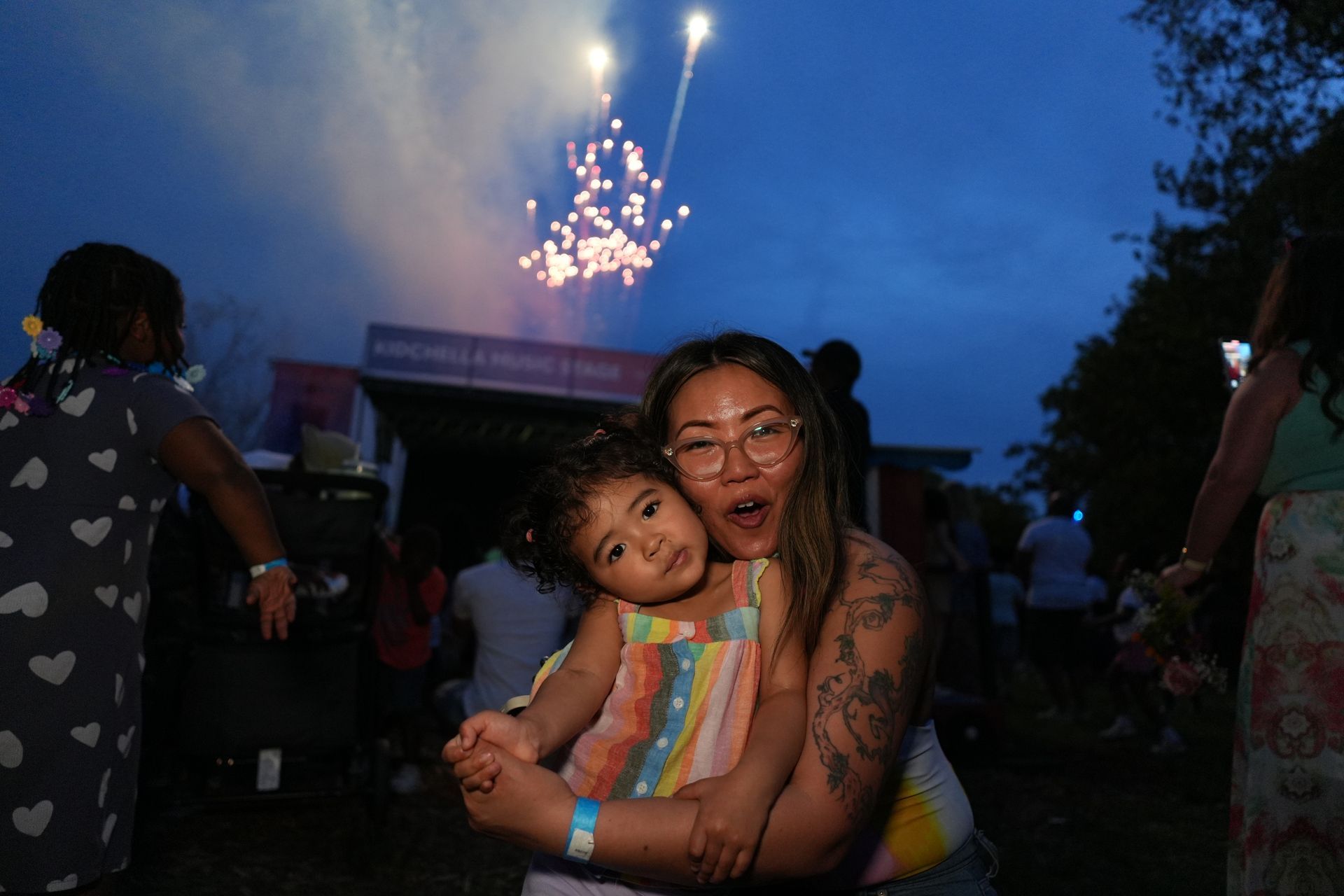 Woman hugging a child outdoors at dusk, smiling as fireworks burst overhead.