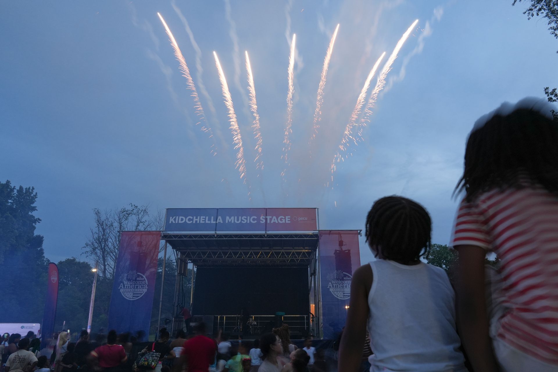 Fireworks over an outdoor concert stage at dusk, with a crowd watching in the foreground.