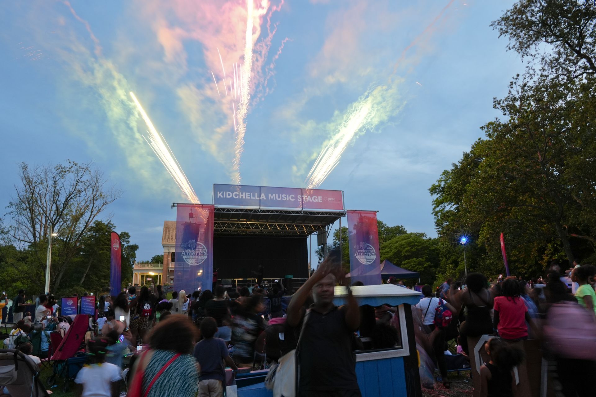 Crowd watching an outdoor stage with fireworks streaking into a colorful evening sky