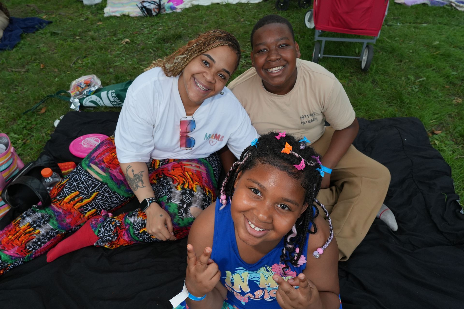 Three people smiling on a blanket at a grassy outdoor gathering, with colorful bags and chairs nearby.