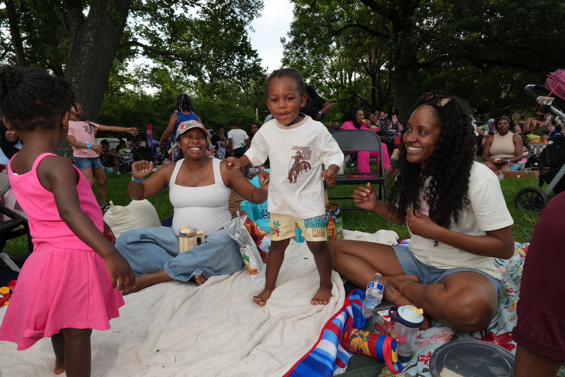 Family sitting on blankets in a park, a toddler standing in the center during an outdoor gathering.