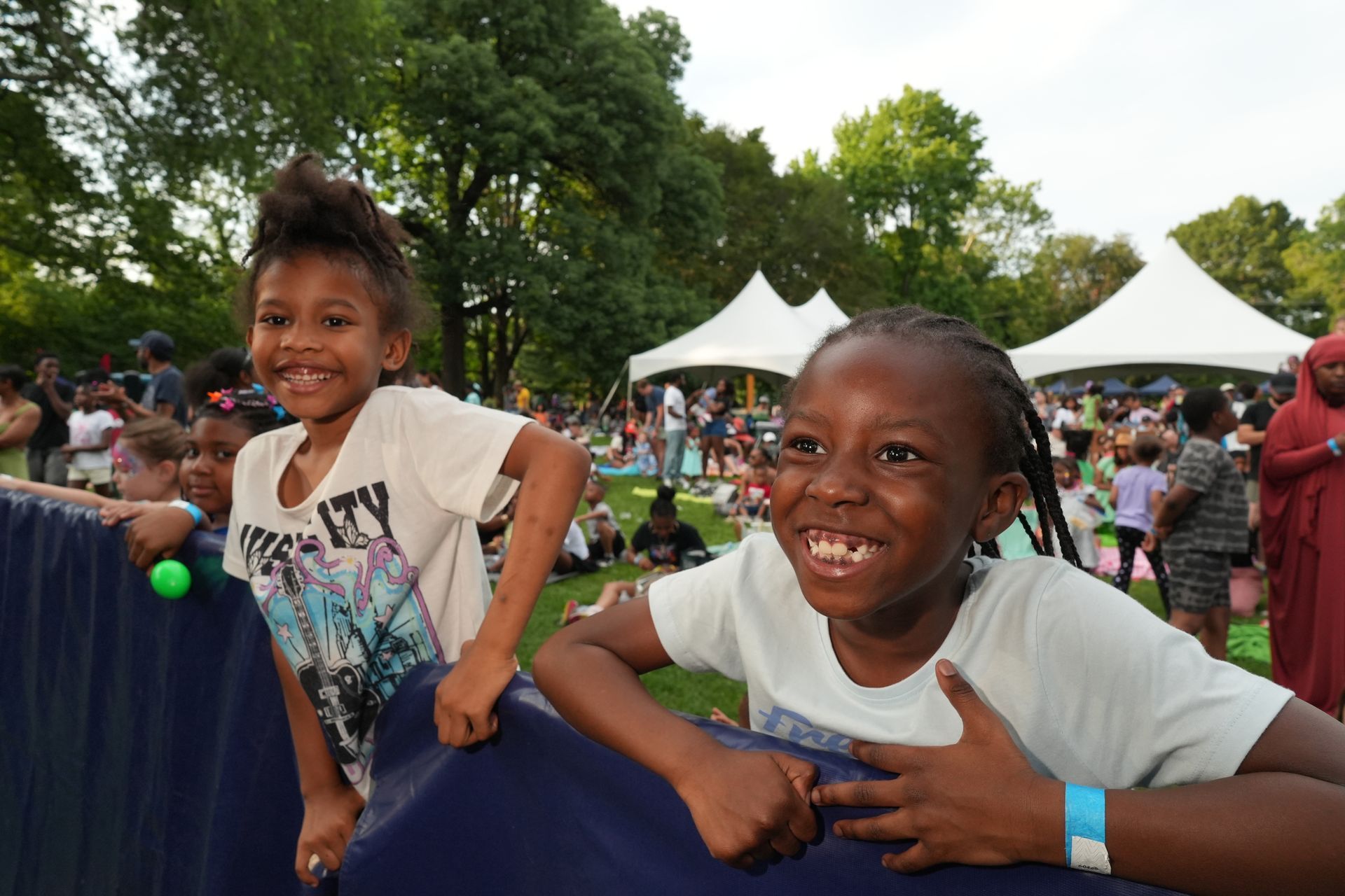 Two smiling children leaning on a blue barrier at an outdoor event with tents and trees.
