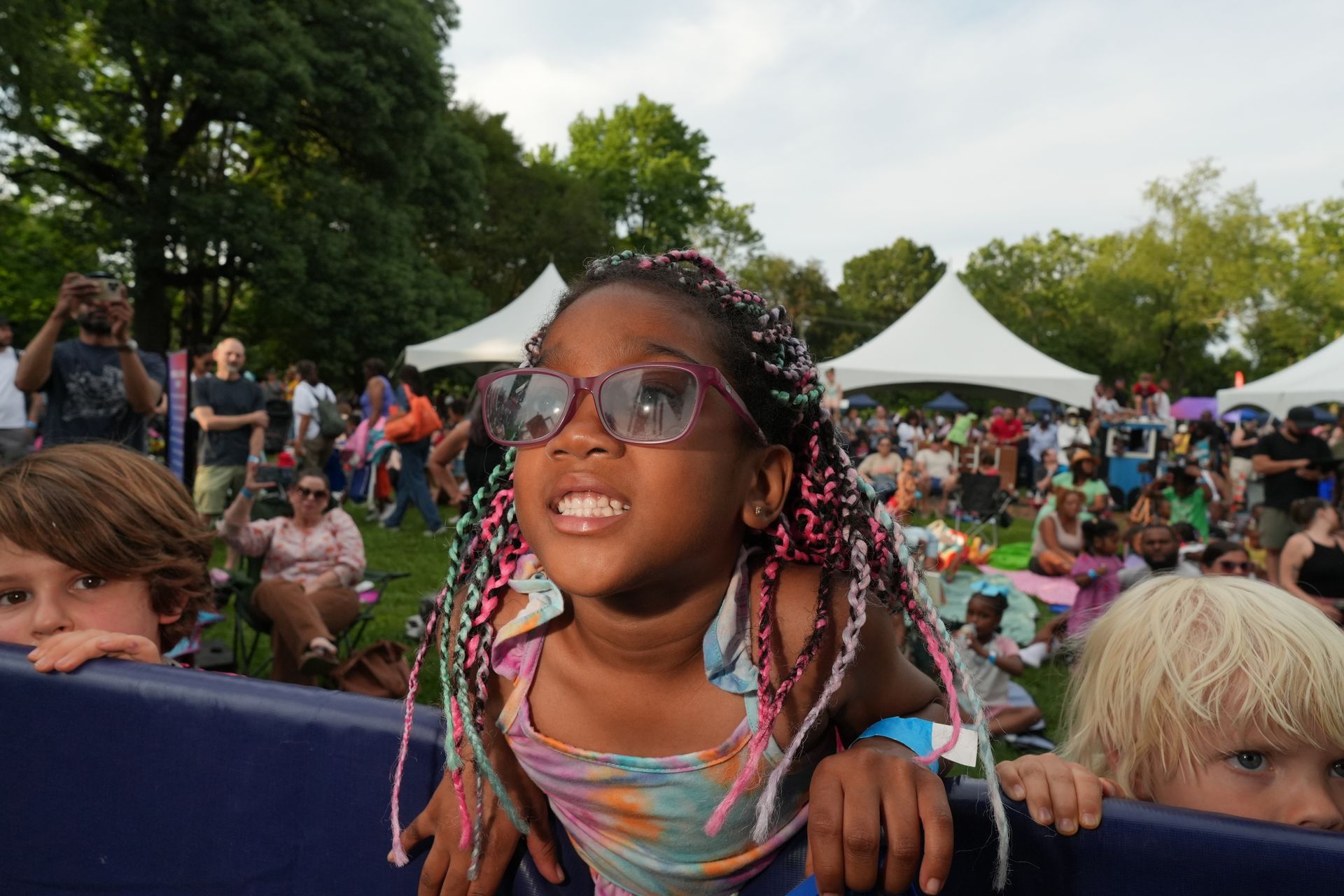 Child in sunglasses leaning on a barrier at an outdoor festival, smiling amid a crowd and tents.