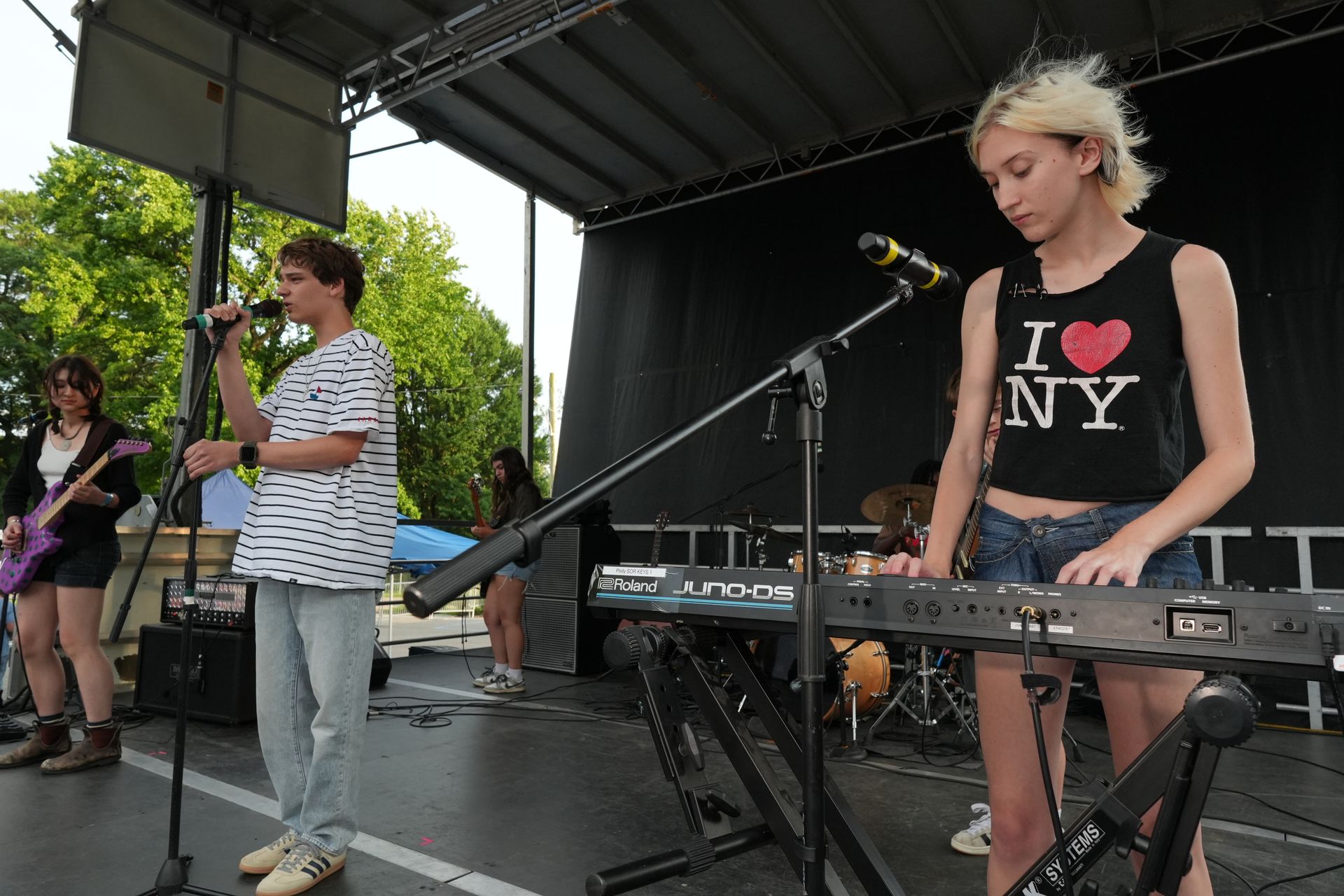 Band performing on an outdoor stage, with a keyboardist in a black I ♥ NY shirt in the foreground.