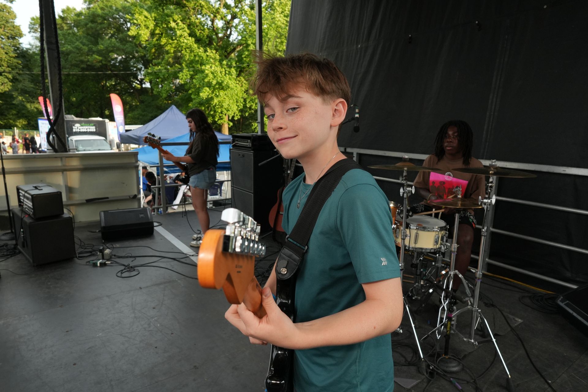 Teen musician playing electric guitar on an outdoor stage, with drummer and sound equipment behind him.