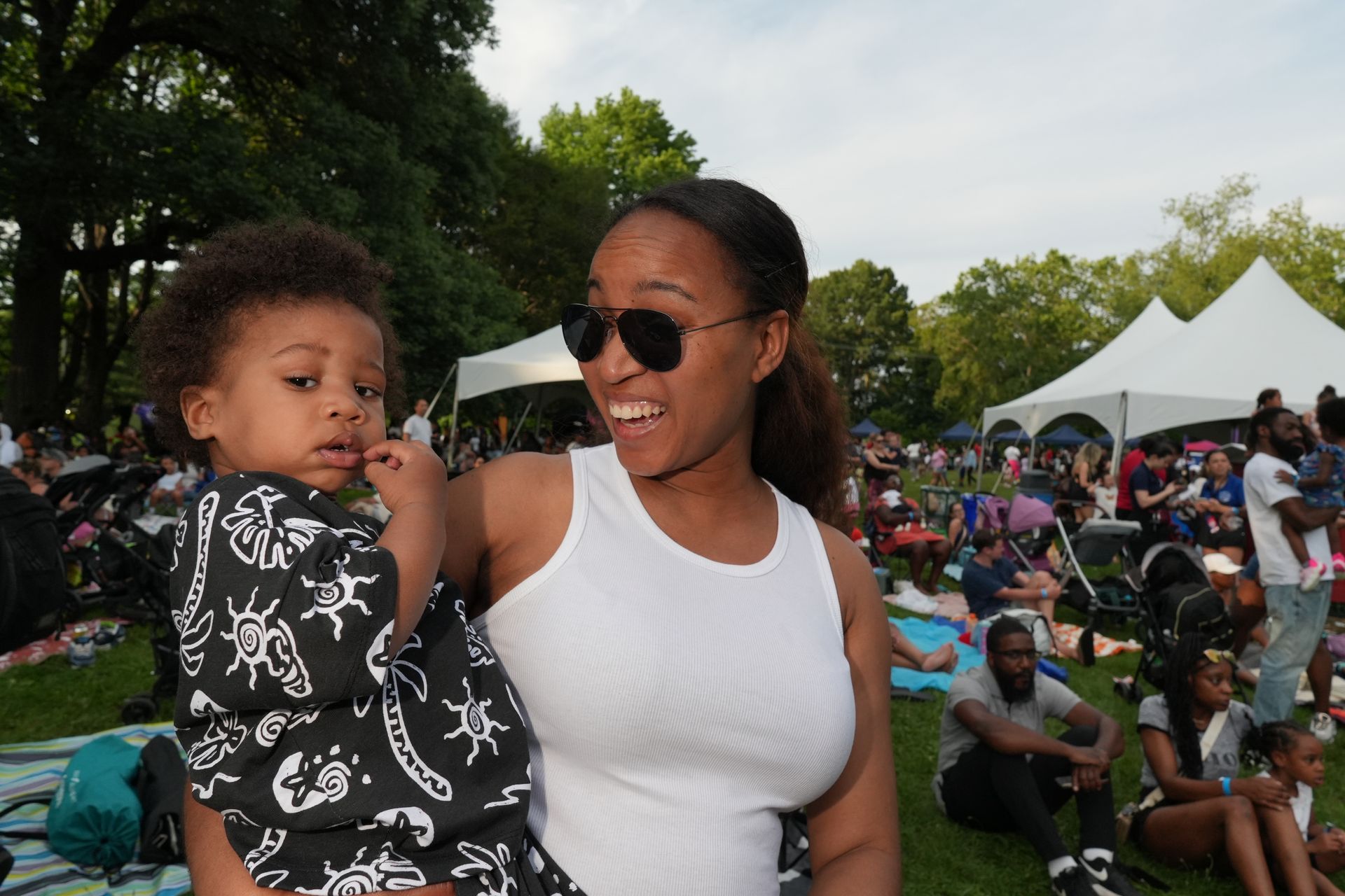 Woman in white tank top holding a child at an outdoor festival with tents and a seated crowd