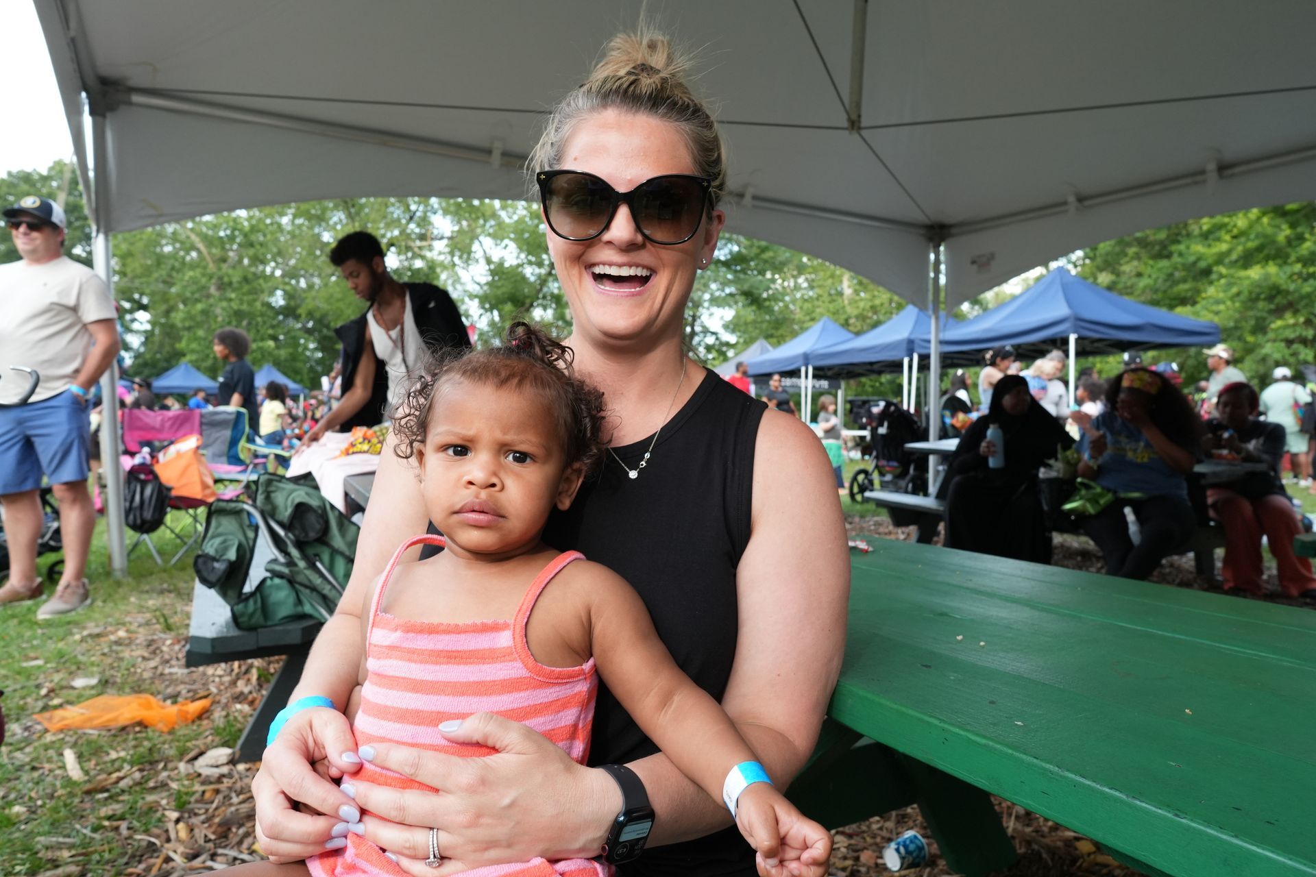 Woman in sunglasses holding a child at an outdoor event under a large white tent