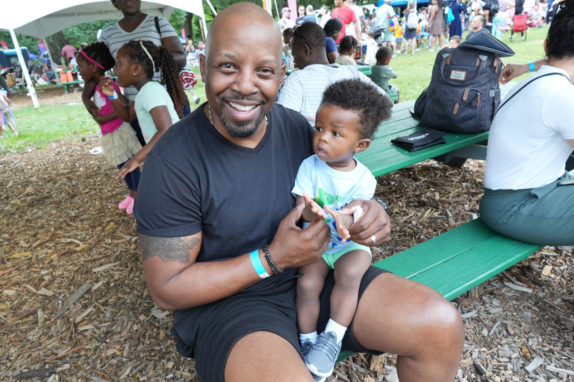 Smiling man holding a toddler on a green picnic bench at an outdoor gathering