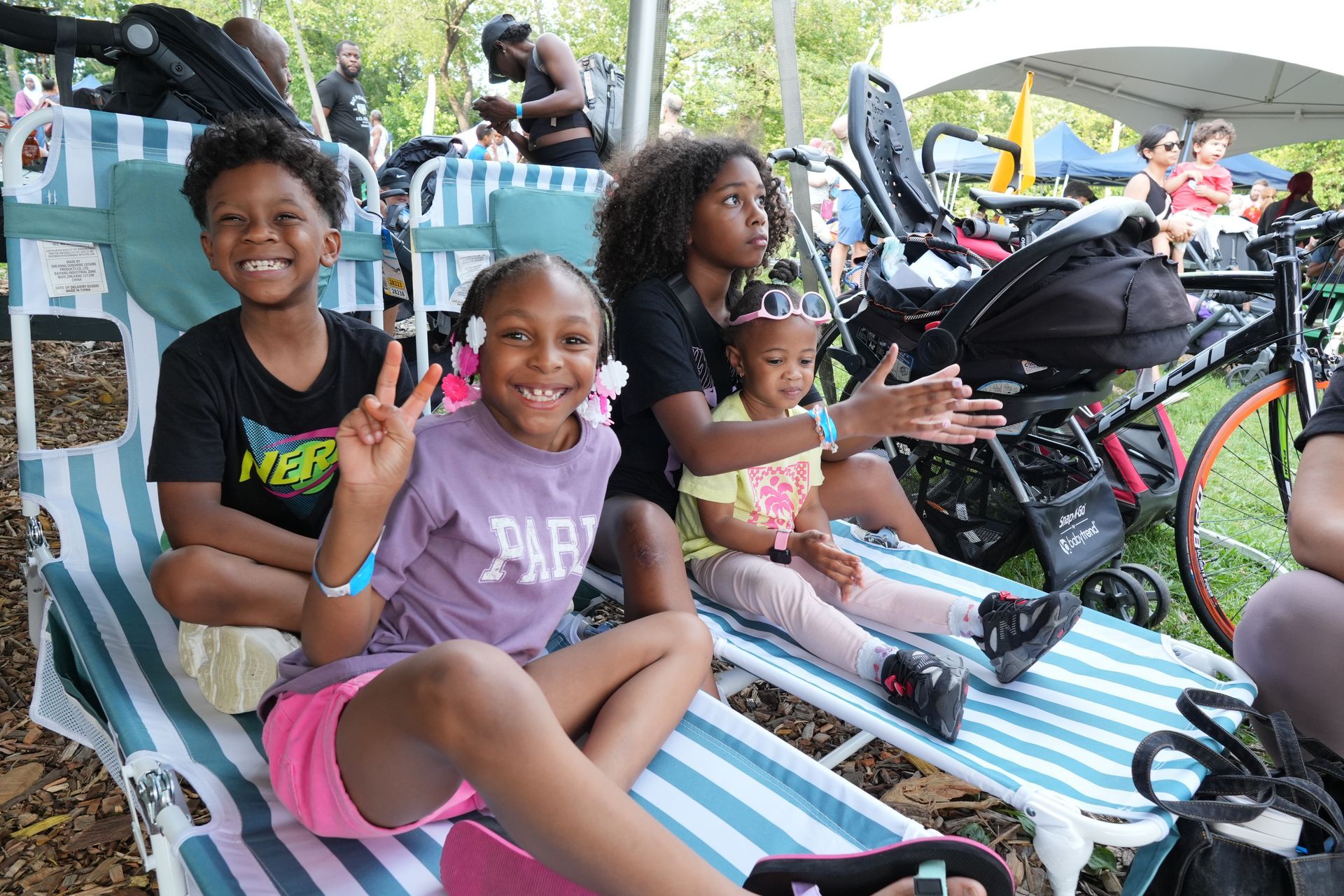 Three smiling people sitting in striped lounge chairs at an outdoor event, waving at the camera.