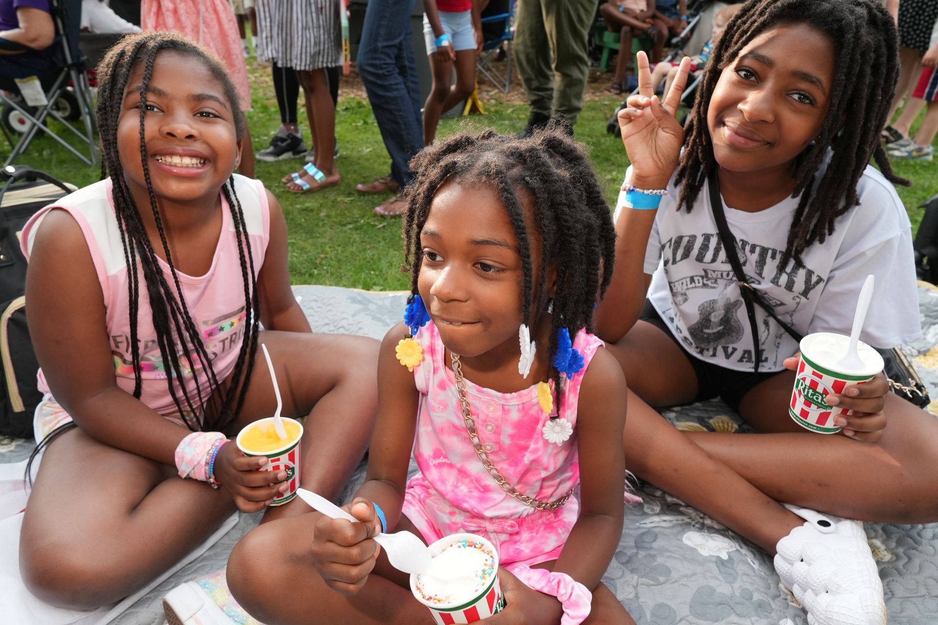 Three children sitting outdoors, smiling and holding cups of dessert on a paved surface