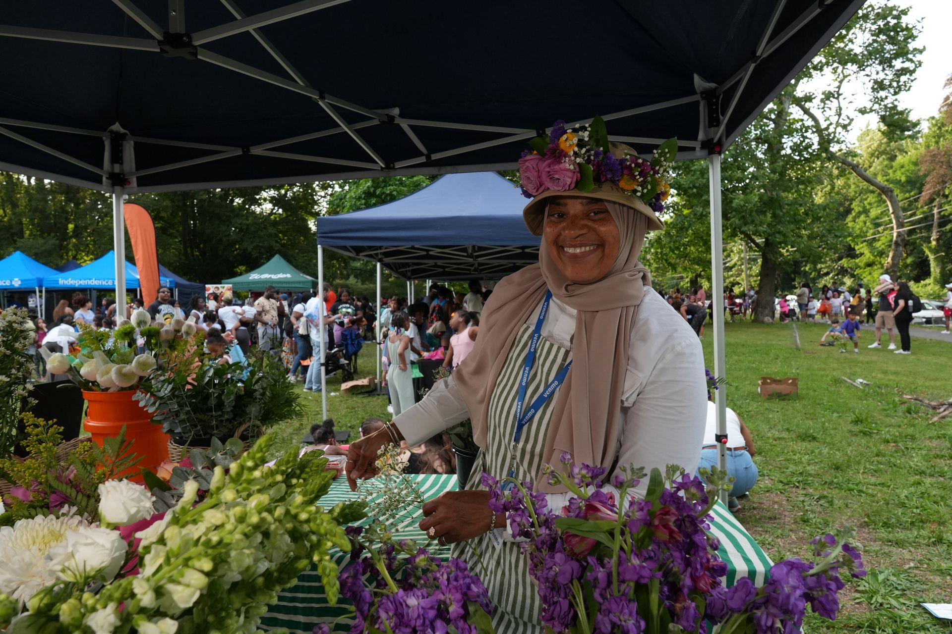 Flower vendor smiling under a canopy at an outdoor market, surrounded by colorful bouquets and tents.
