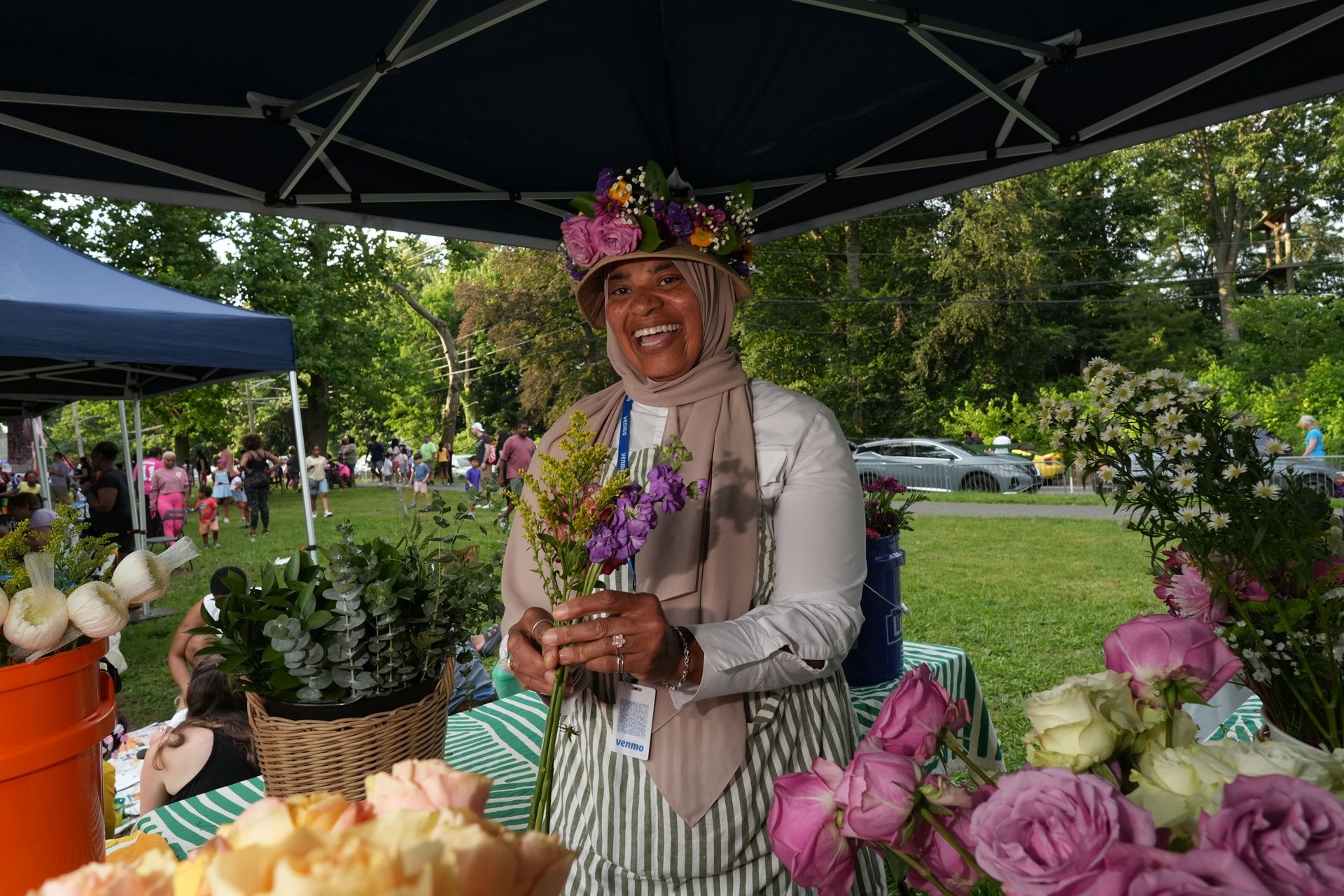 Flower vendor smiling under a canopy at an outdoor market, surrounded by colorful bouquets and potted plants.