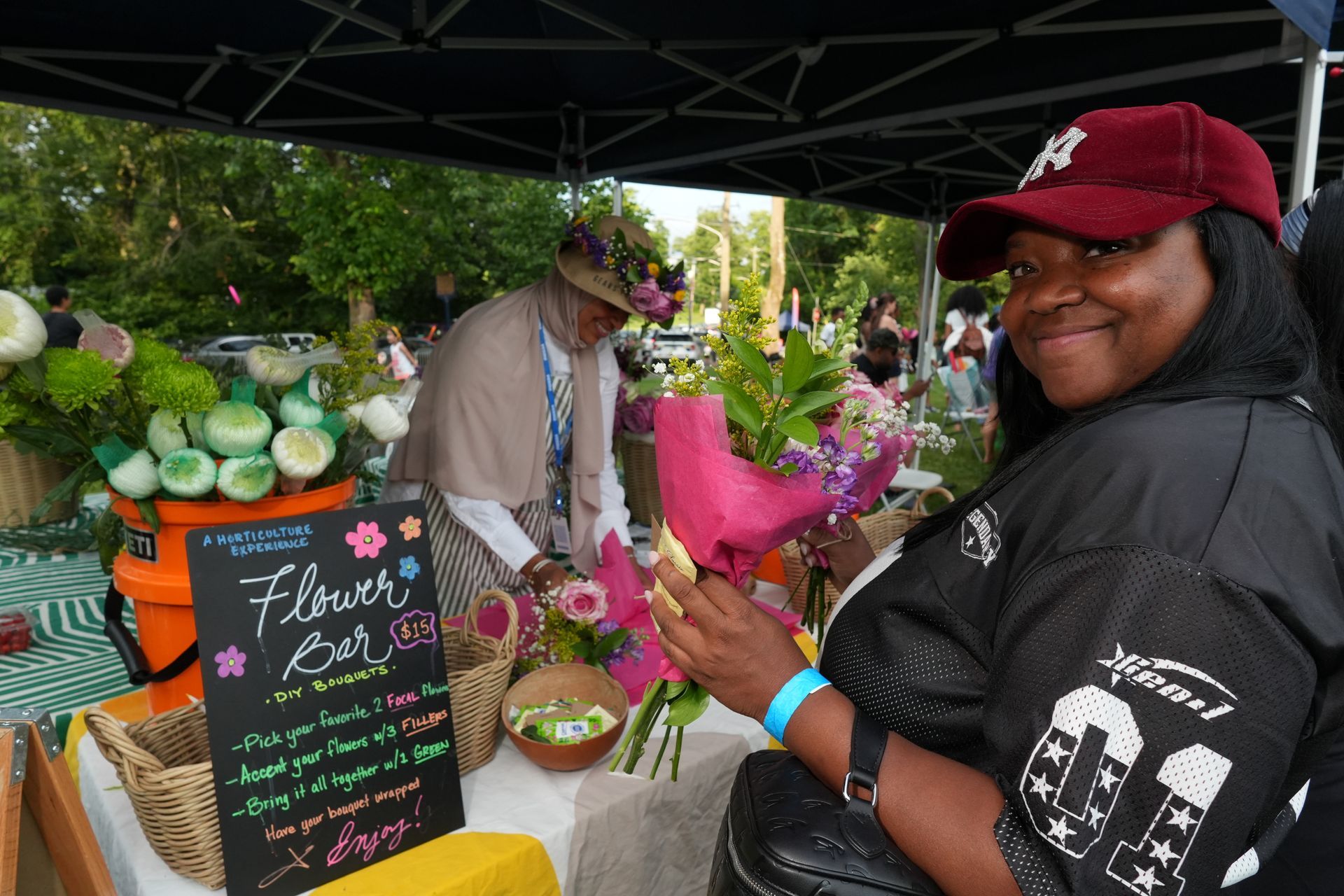 Person smiling at a flower market stall, holding a pink bouquet under a canopy.
