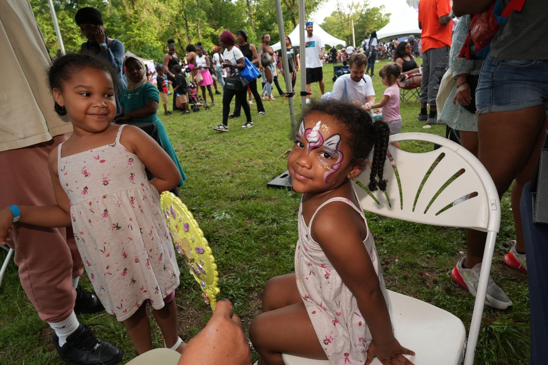 Children at an outdoor gathering, one seated smiling with face paint, others playing on the grass